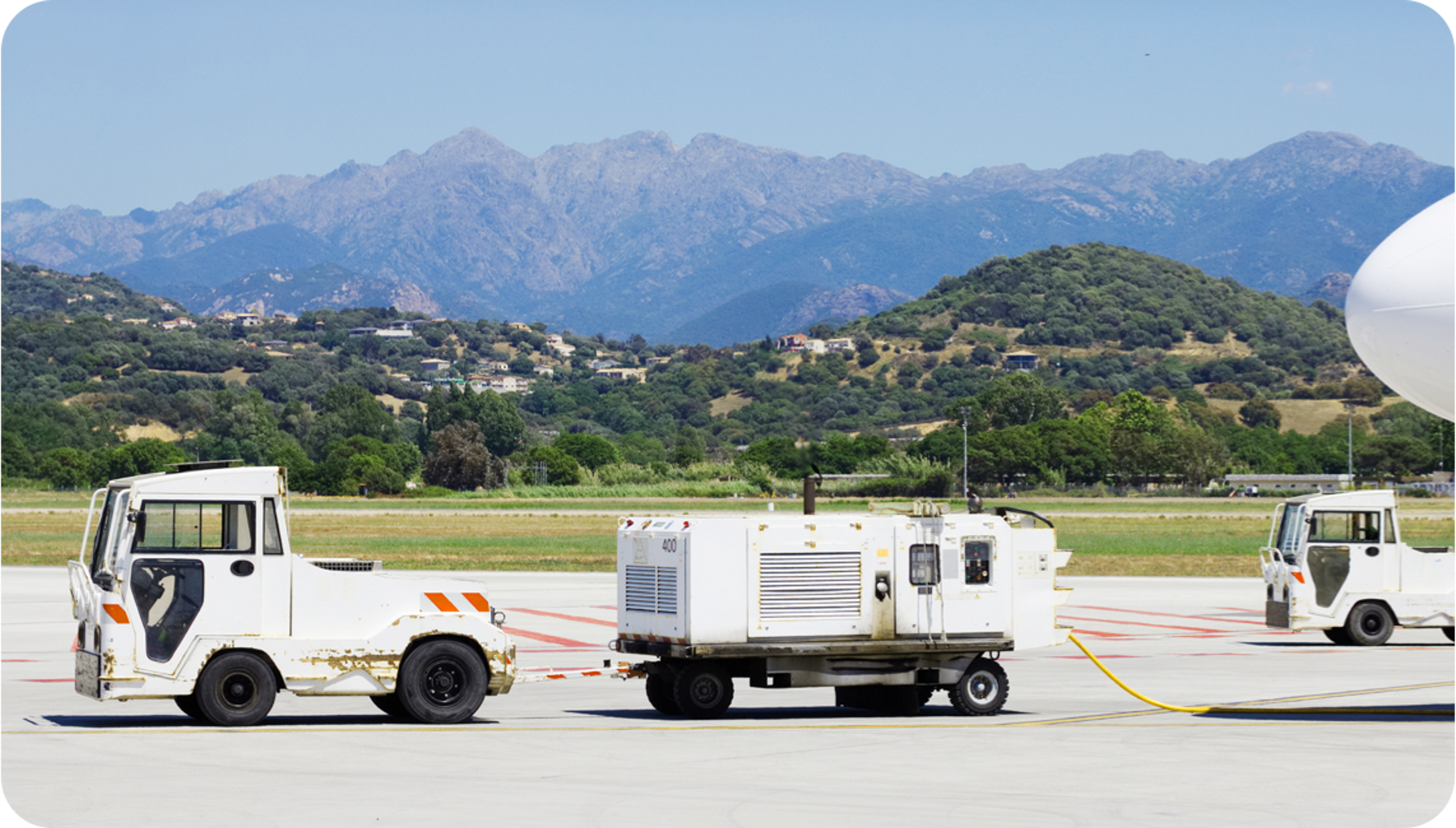 Pictire of white truck and white battery trailer at an airport runway with trees and mountains in the horizon