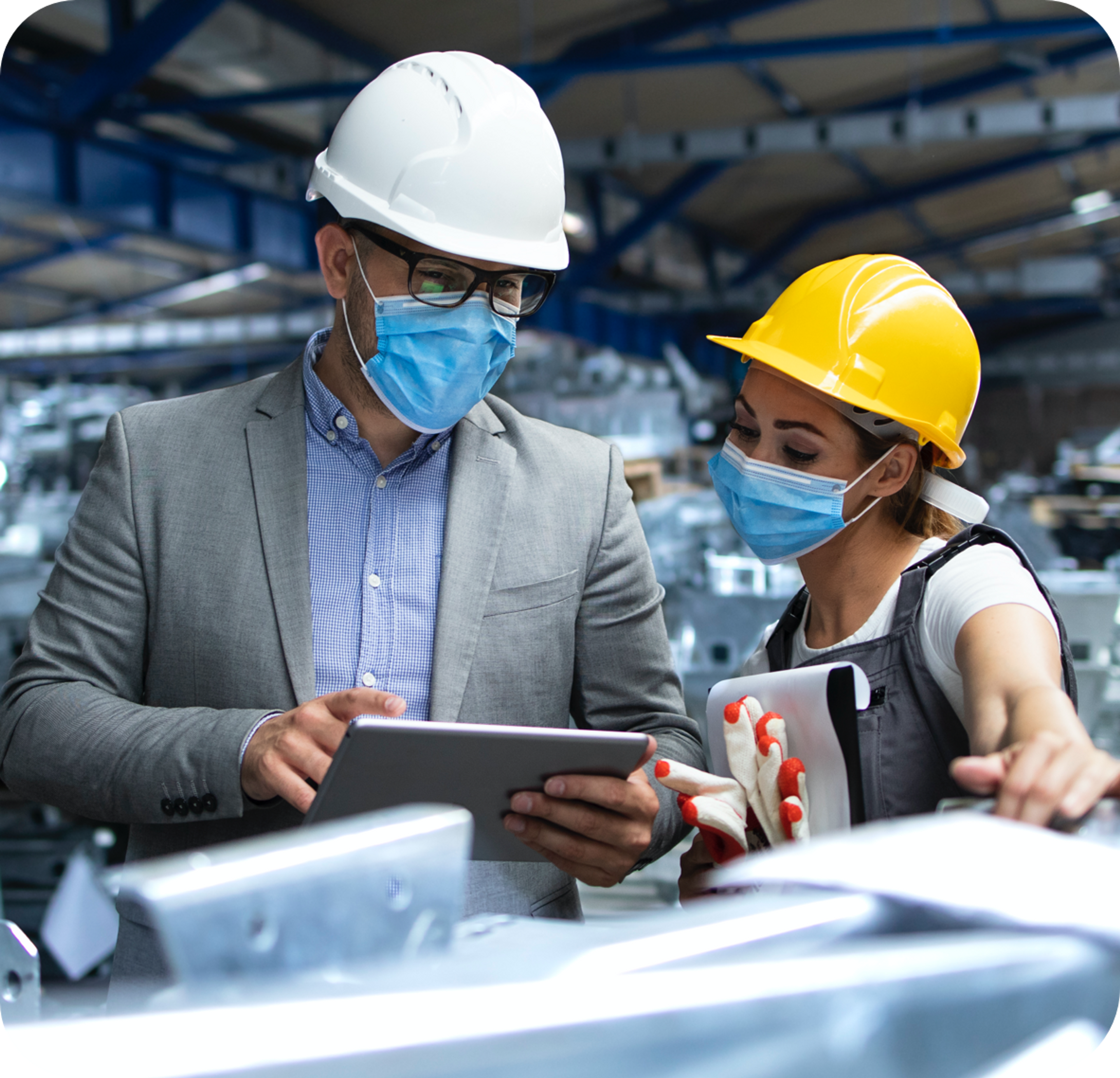 Picture of Caucasian Man in a warehouse setting wearing white hard hat and blue surgical mask and gray suit jacket and blue collared shrirt talks to an ethnic woman wearing jean coveralls, white tshirt and yellow hard hat the contents shown in his Ipad