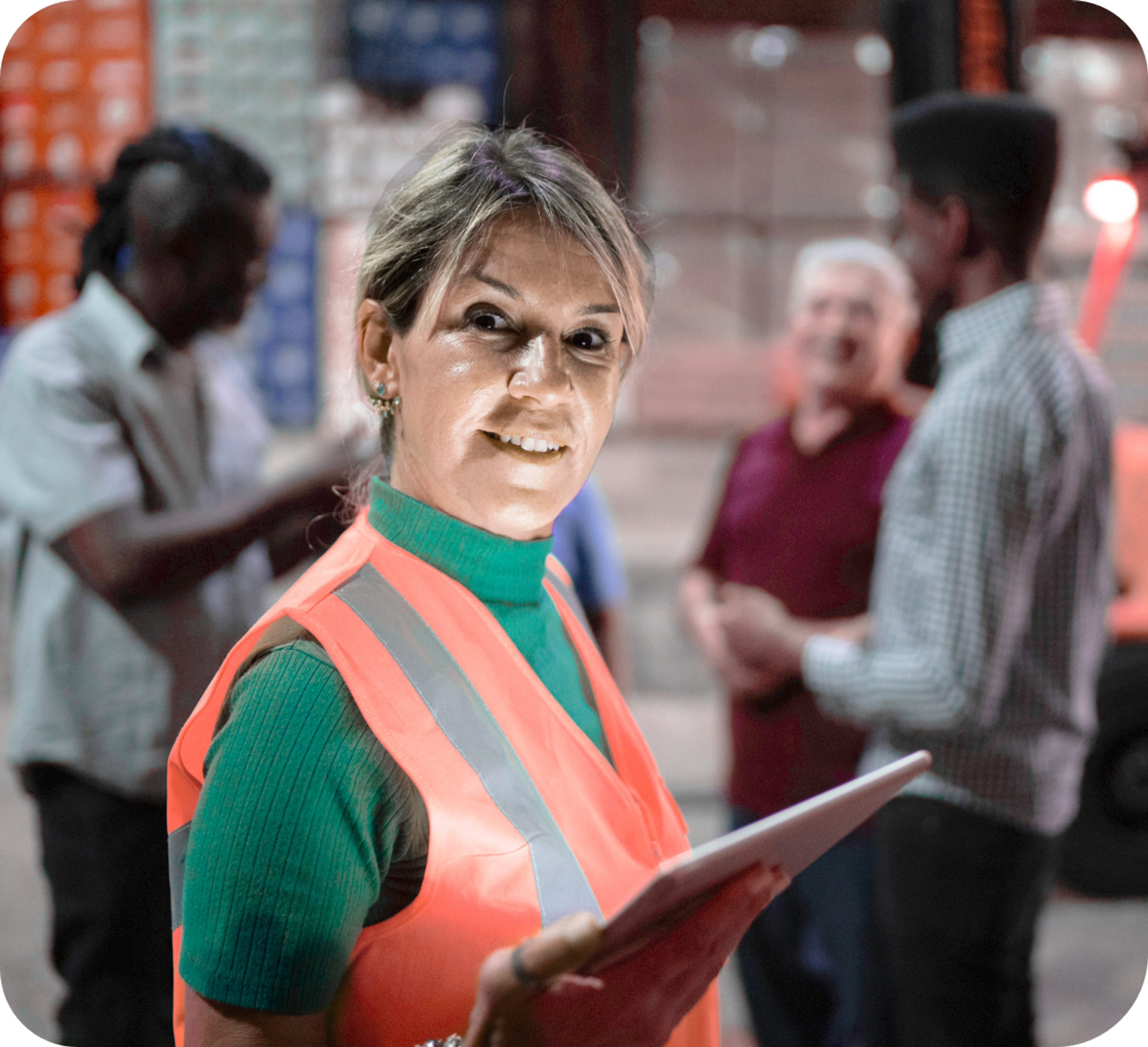 Caucasian middle aged woman in a warehouse wearing a green turtle neck and orange safety vest smiling and holding an Ipad while three men are blurred in the background