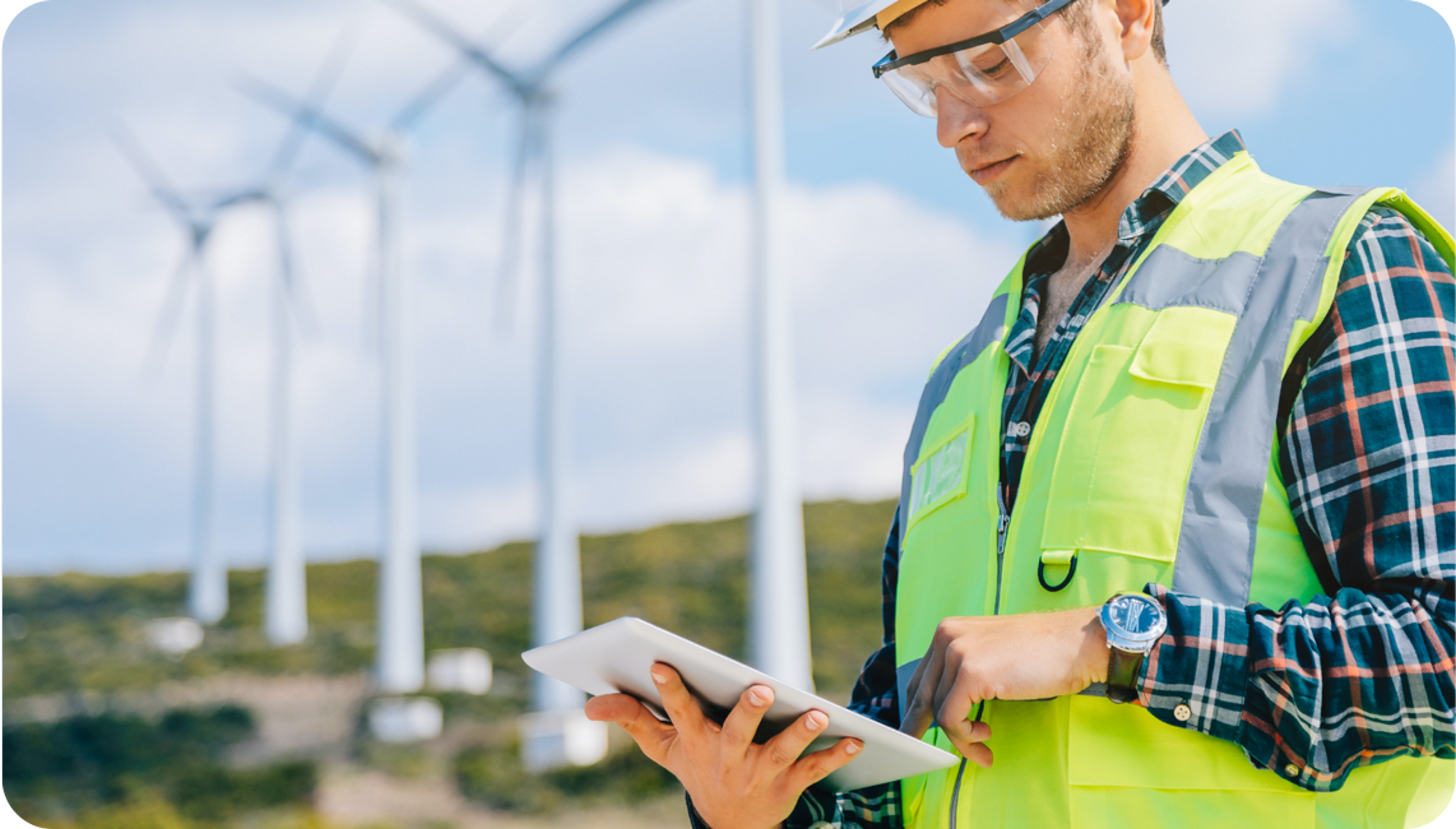 Picture of caucasian man wearing safety glasses and yellow safety vest typing on an iPad and three wind turbines spinning in the background 