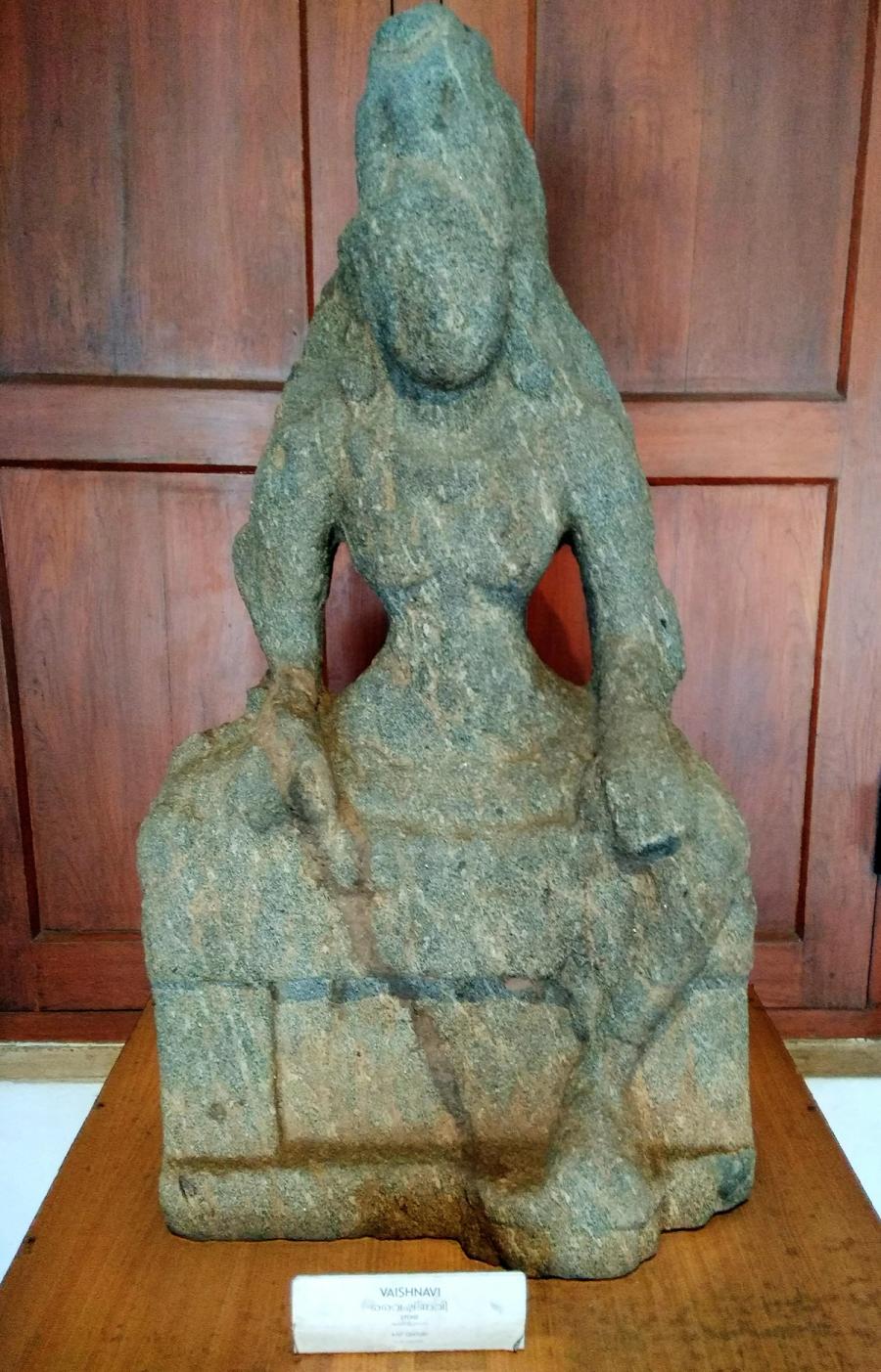 Weathered stone sculpture of the goddess Vaishnavi, museum-labelled, seated.