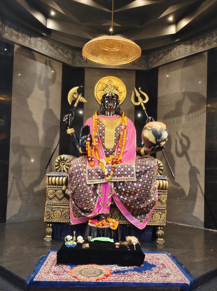 A dark-complexioned idol of Shani Dev, seated on a throne holding a trident and axe, at Shani Dham, Char Dham Vrindavan