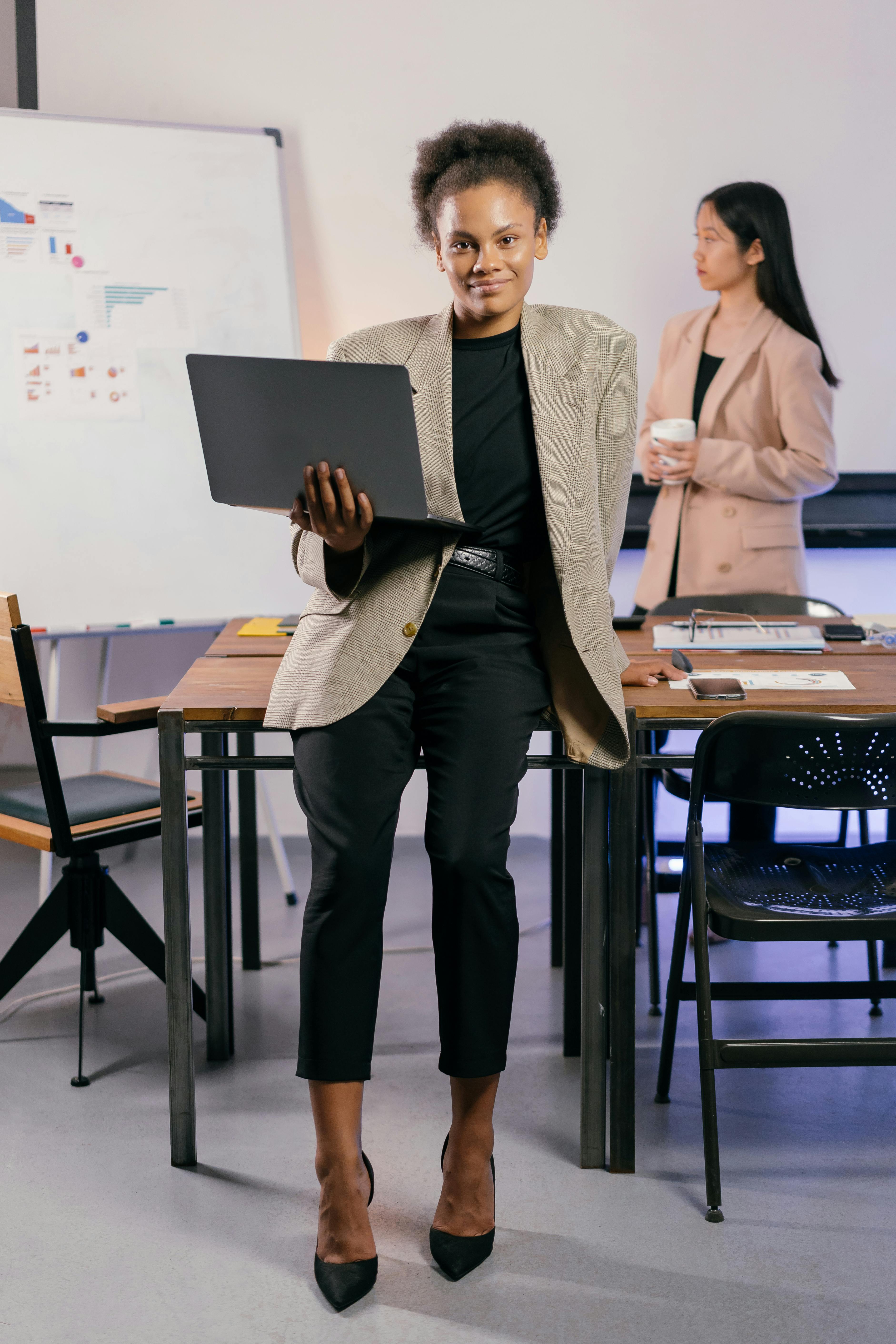 Professional woman sitting on edge of desk holding laptop.