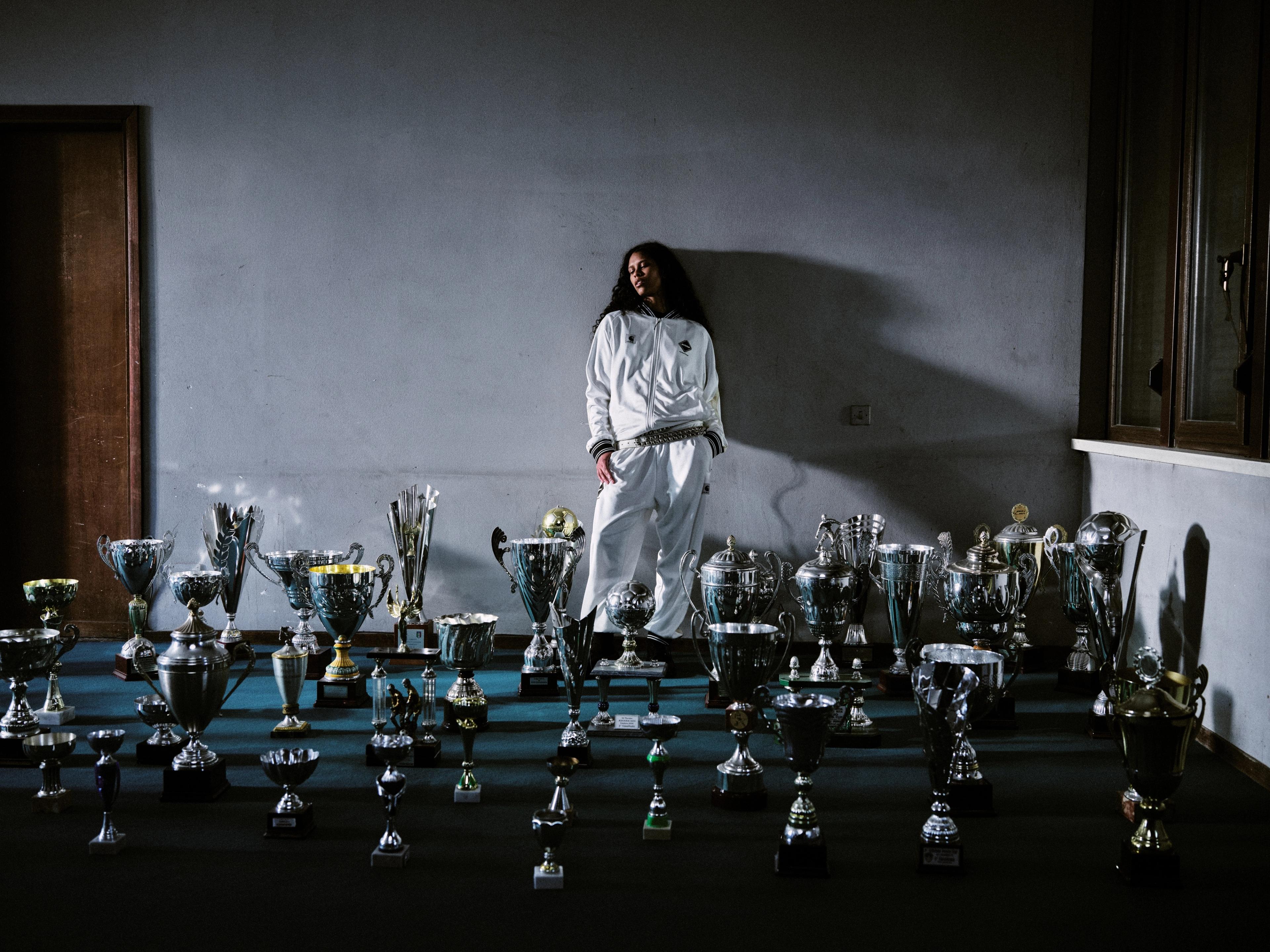 A person in a white Carhartt WIP tracksuit stands against a dimly lit wall behind a large collection of shiny trophies arranged on the floor.