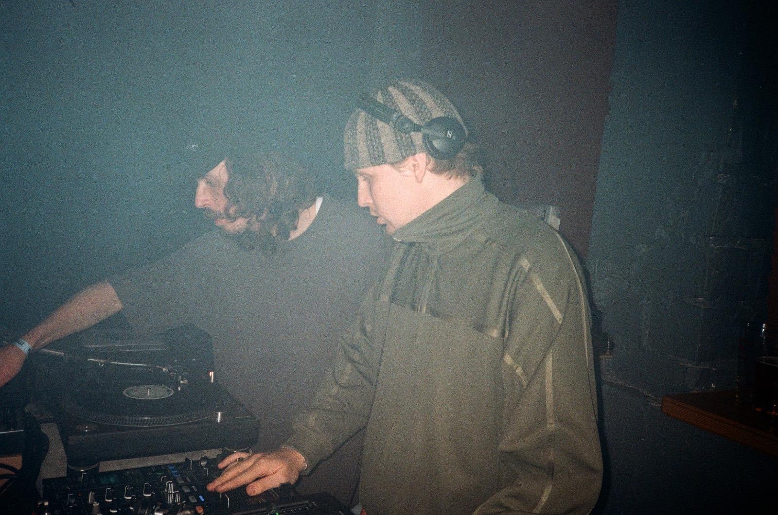 Two DJs stand side by side in a dim, smoky club, focused on mixing tracks on turntables and a mixer, with no visible Carhartt WIP branding.