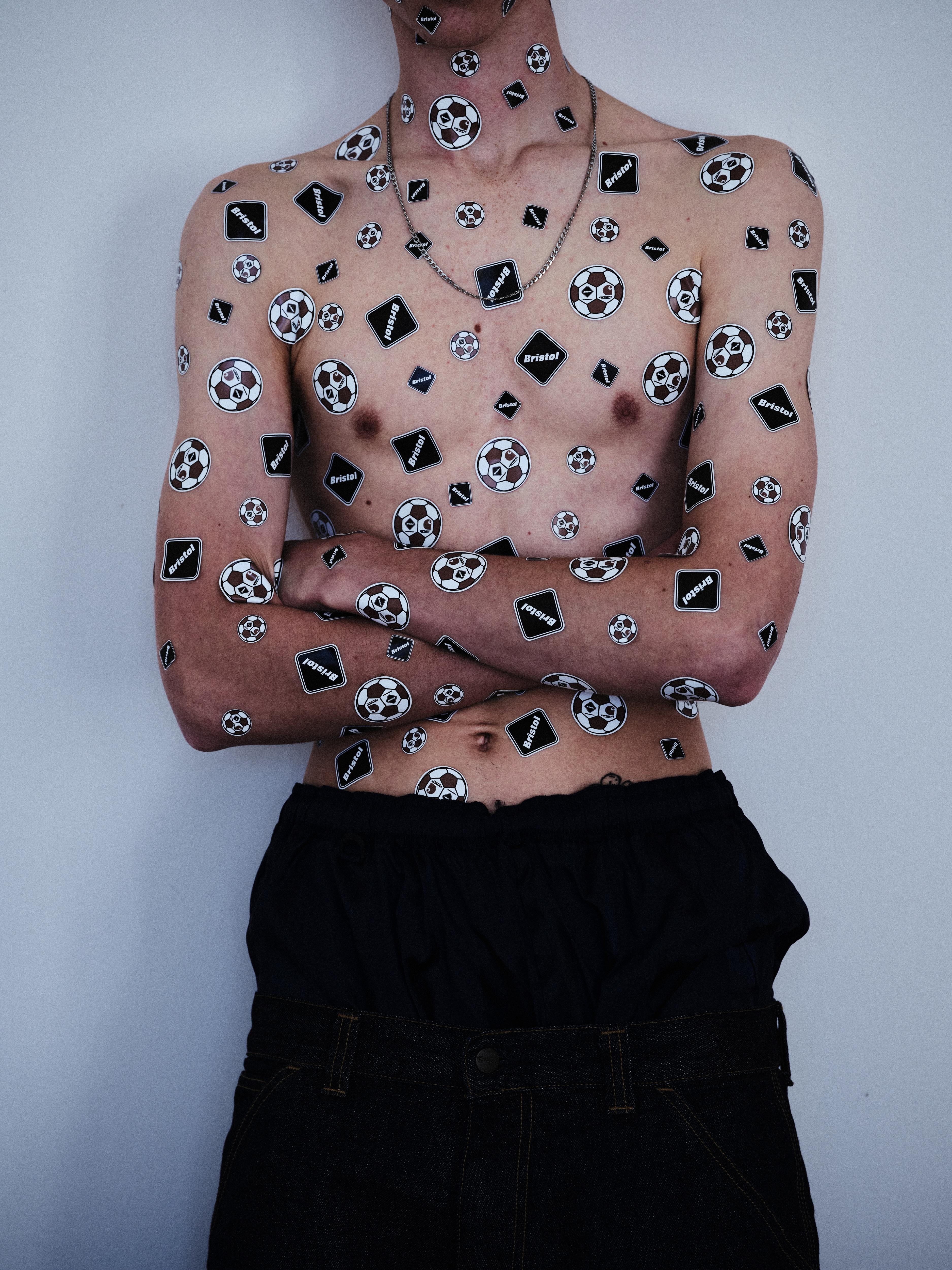 A shirtless person wearing dark pants stands against a plain background with their arms crossed, their torso and arms covered in numerous black-and-white stickers featuring soccer balls and the word “Bristol,” with no visible Carhartt WIP branding.