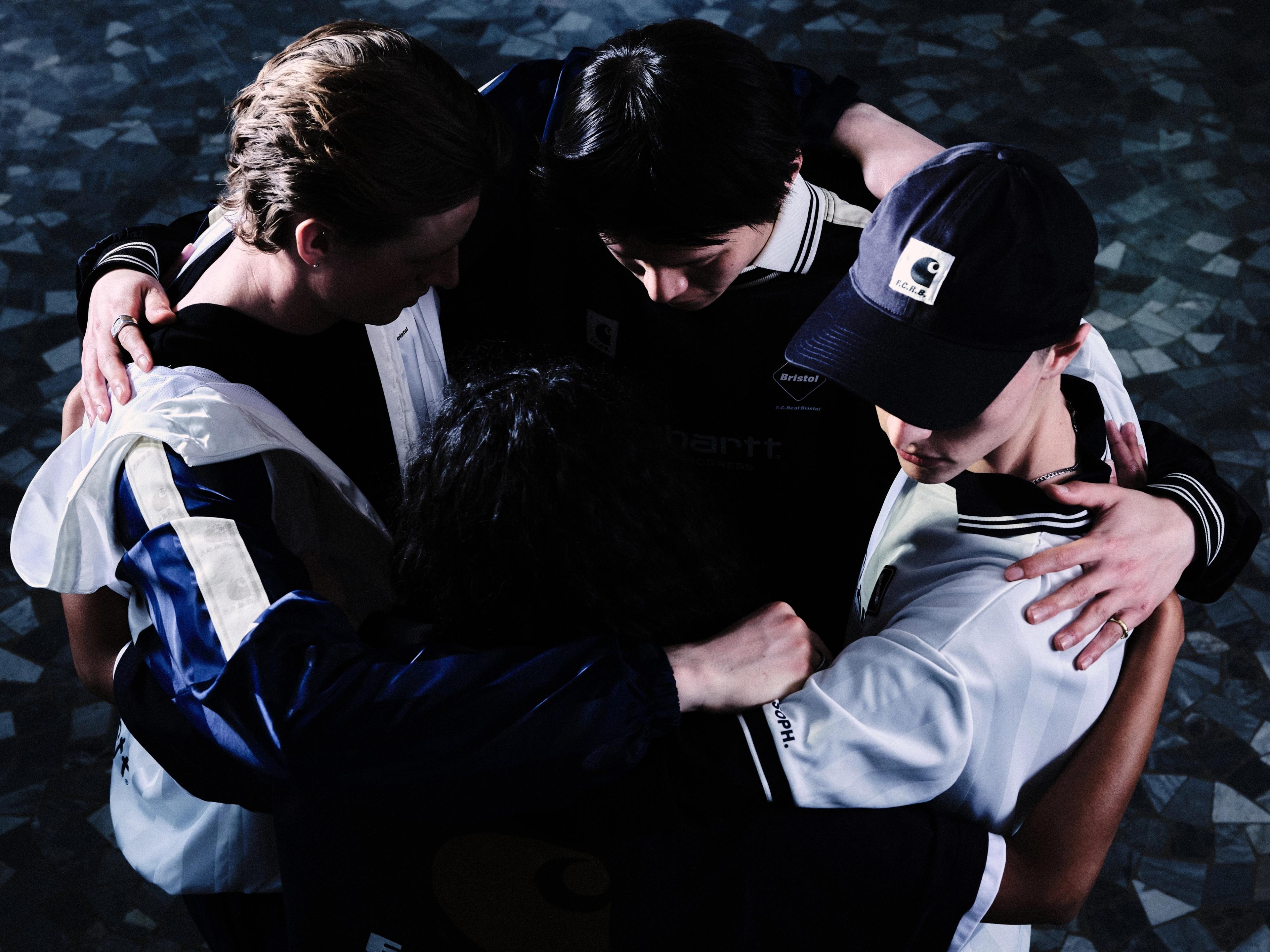 Four people wearing sportswear huddle closely together with arms around each other, one of them wearing a navy cap featuring the Carhartt WIP logo.