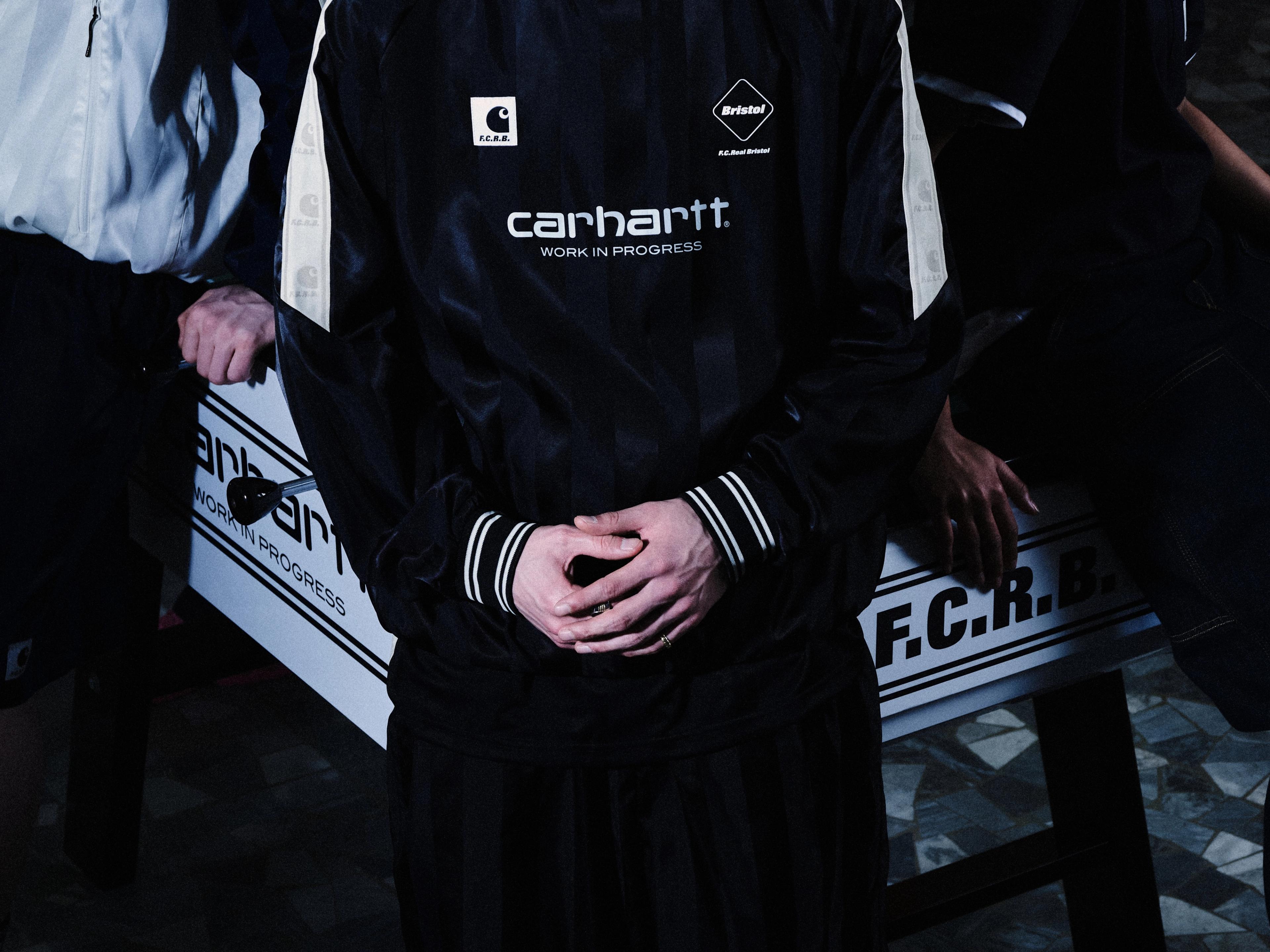 A person stands with hands clasped in front, wearing a black Carhartt WIP track jacket with visible branding, next to a foosball table in a dimly lit setting.
