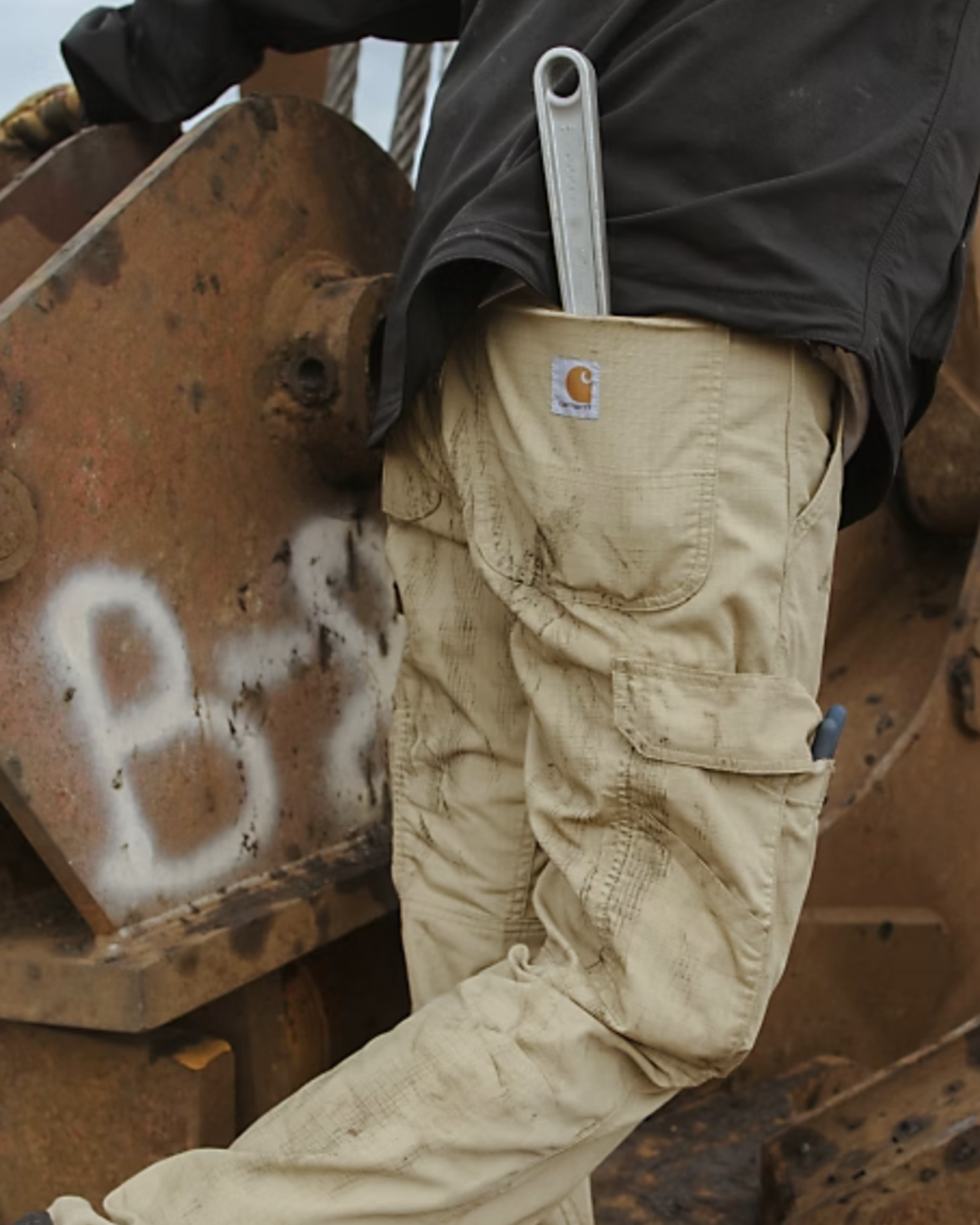 A person wearing beige work pants with a wrench tucked into the back pocket stands next to a large, rusted metal structure. The pants are slightly dirty, indicating a work environment.