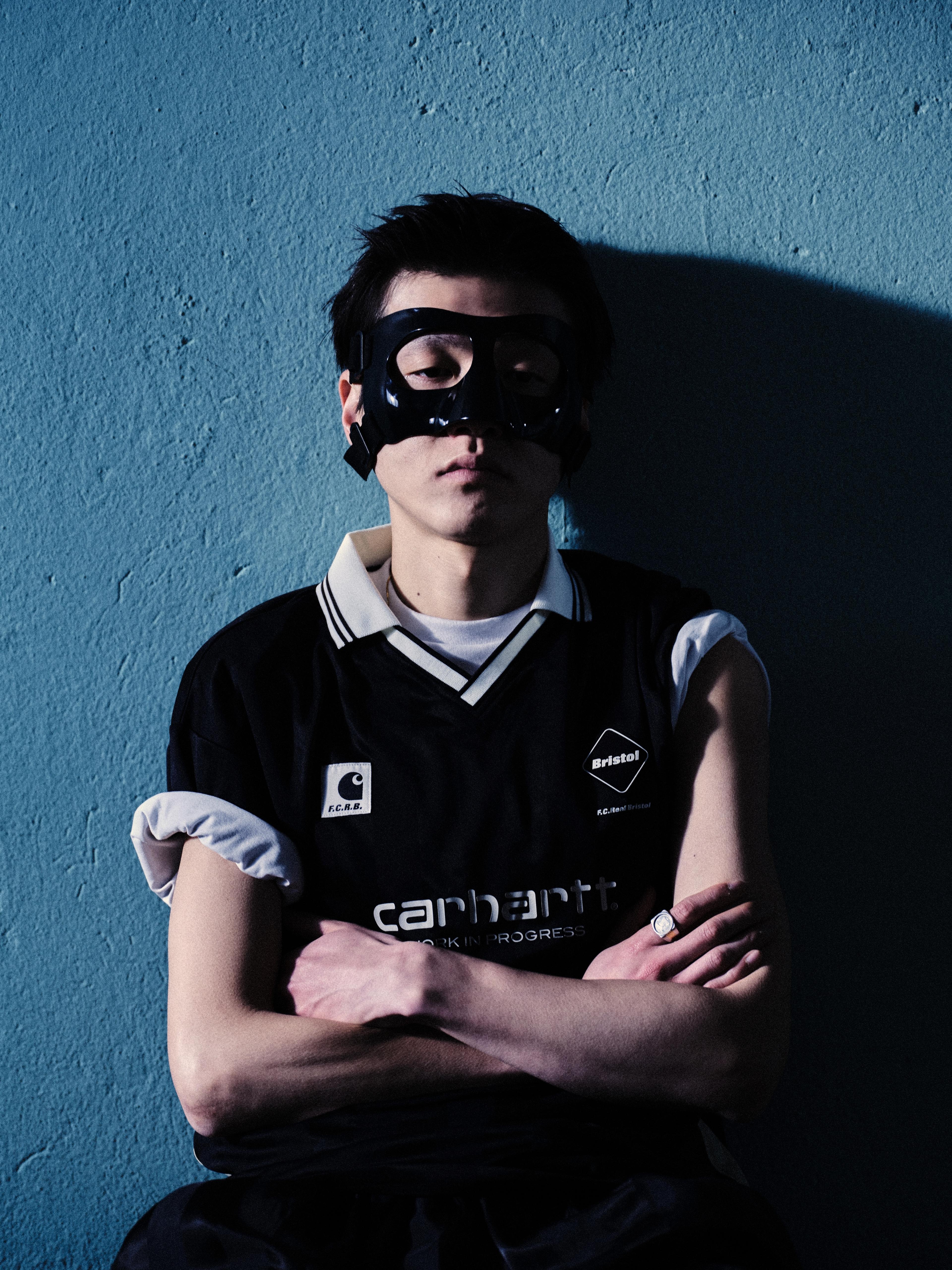 A person wearing a black eye mask and a black sleeveless Carhartt WIP sports jersey stands against a blue wall with arms crossed.