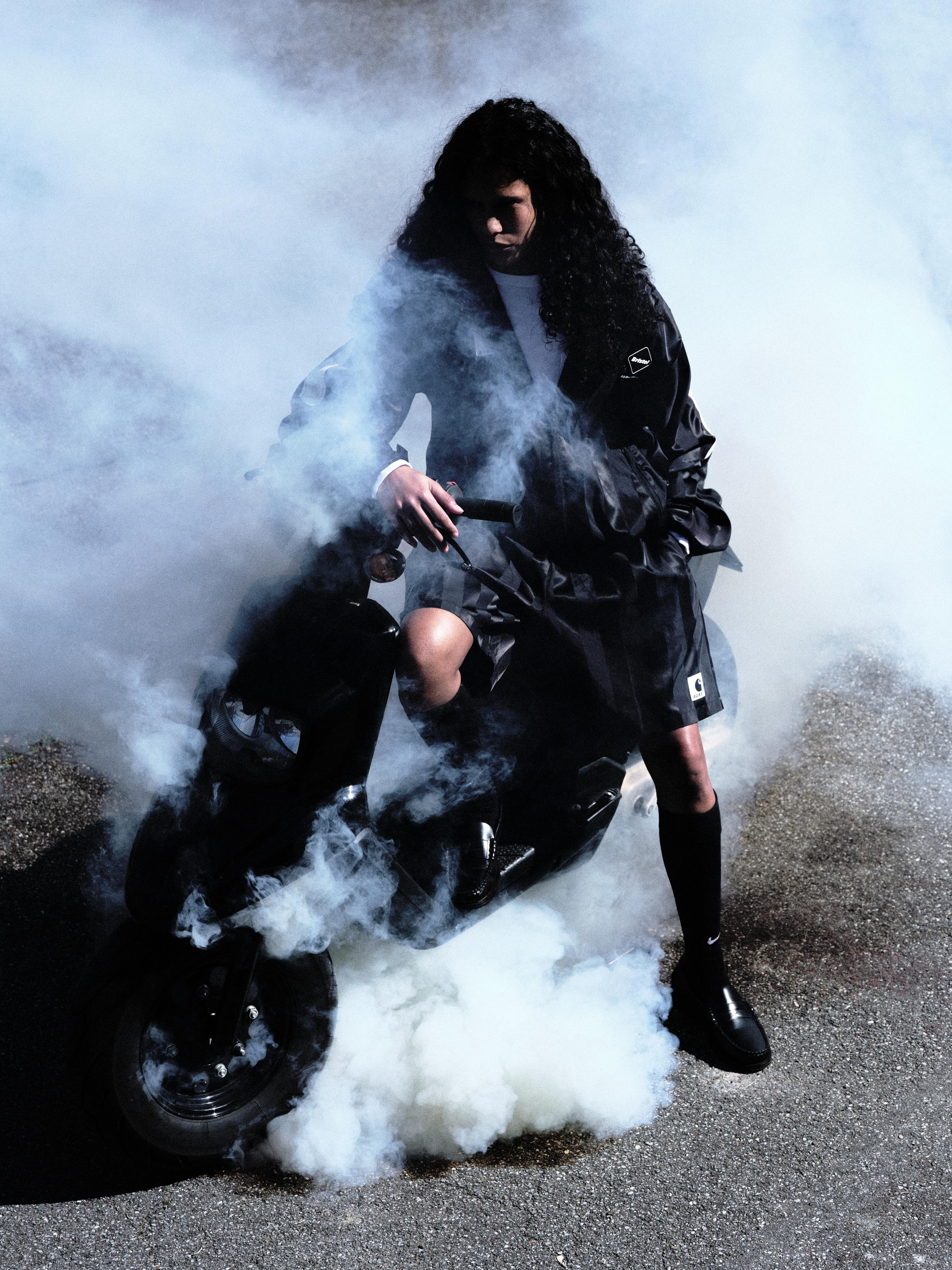 A person wearing a dark Carhartt WIP outfit rides a scooter doing a burnout, surrounded by thick white smoke on an asphalt surface.