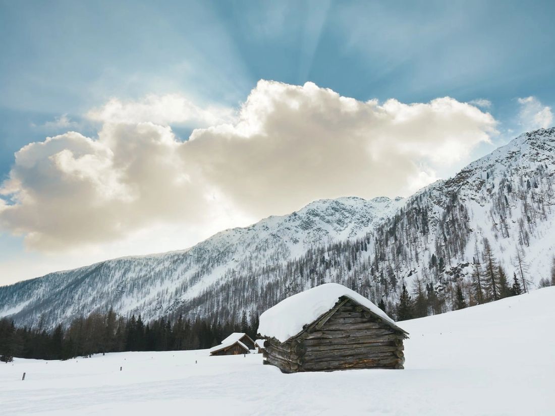 A snow-covered log cabin in an open alpine meadow with sunlight breaking through clouds