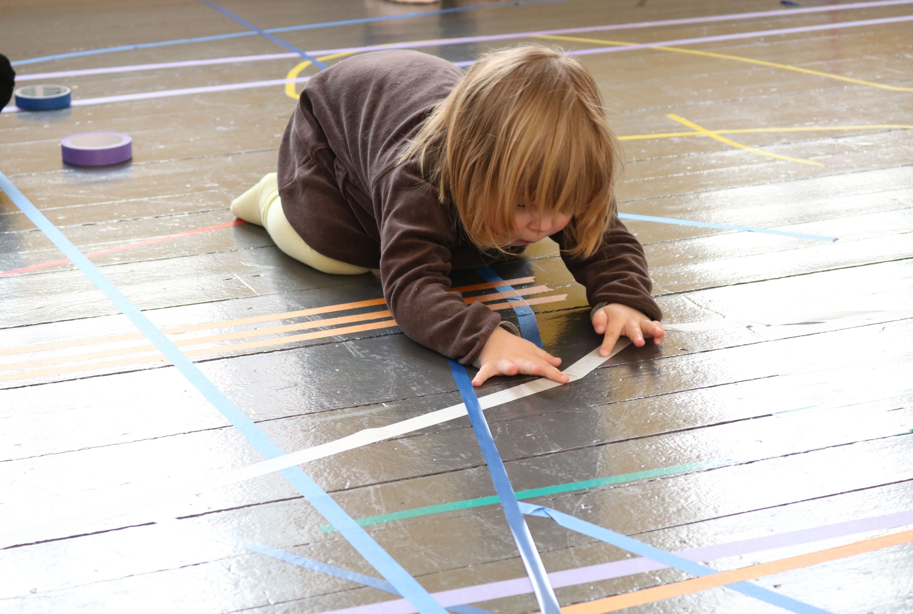 Barn ligger på kne på gulvet og fester teip i ulike farger på gulvet