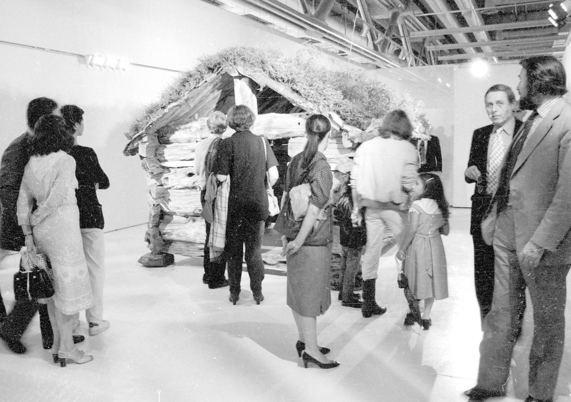 Svart/hvitt-bilde av et lite trebygg på utstilling i museum med tilskuere
