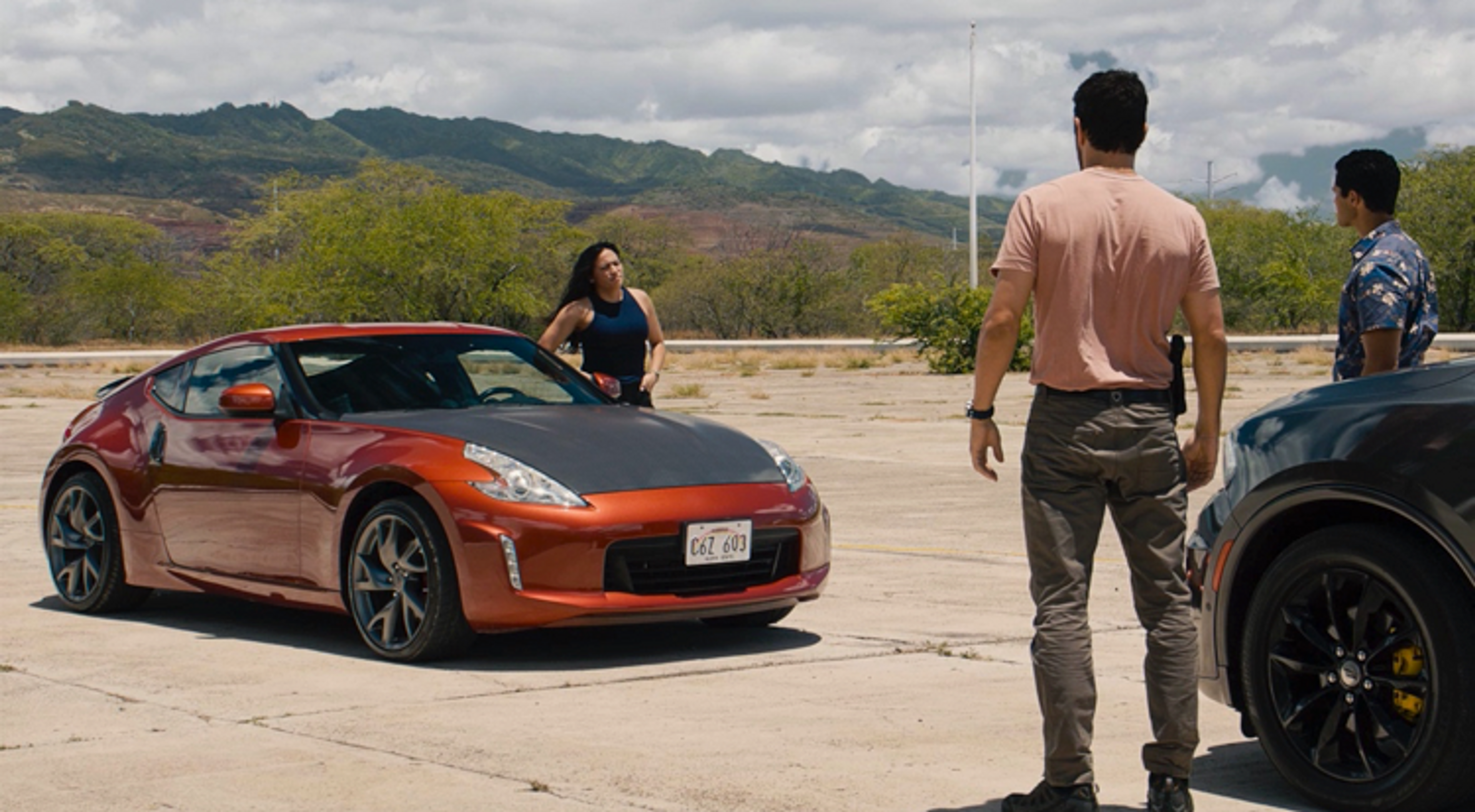 A shot on the set of NCIS. A woman is standing next to an orange Nissan Skyline with a carbon fiber hood while talking to two men who are standing next to a black vehicle.