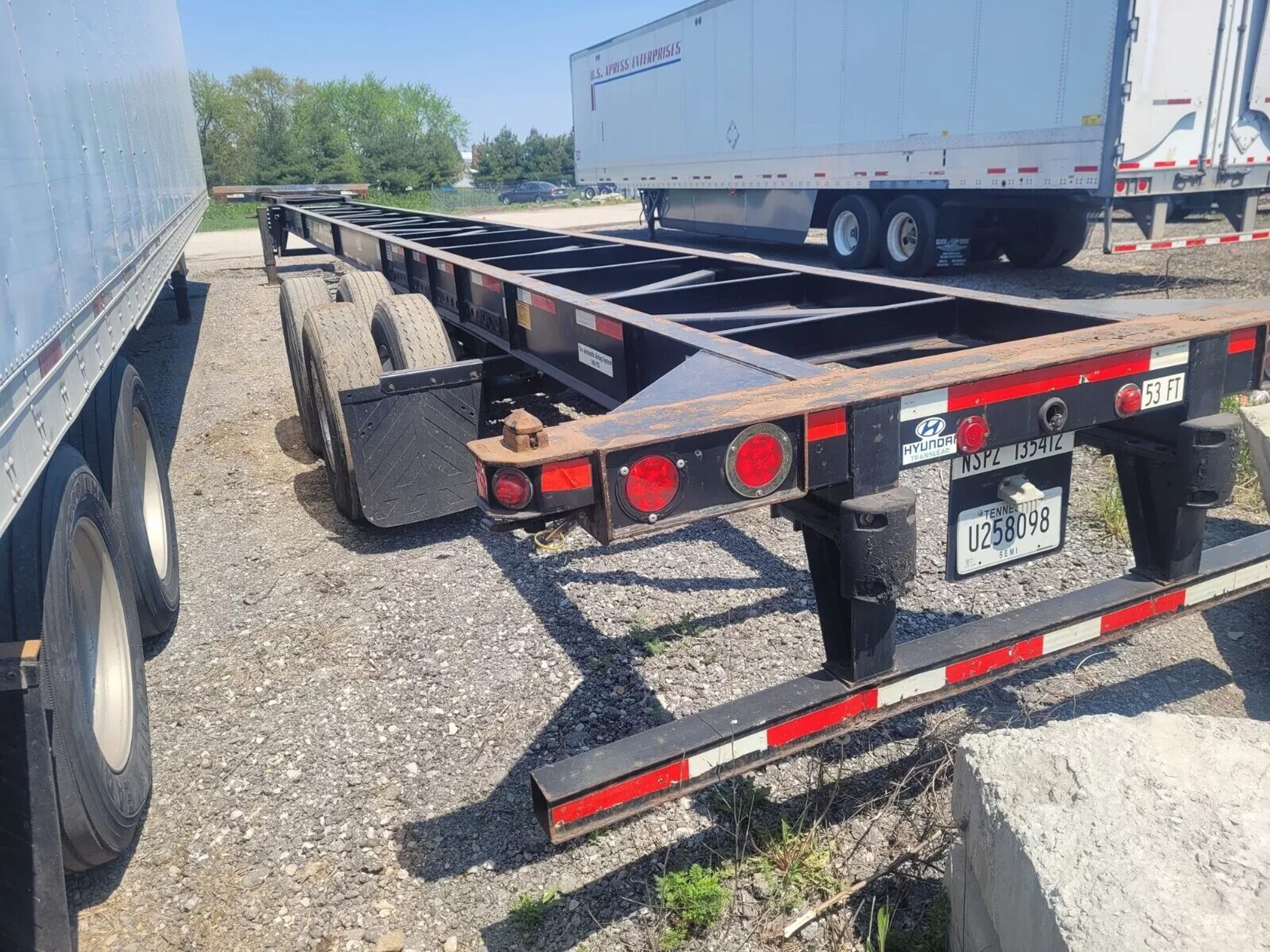A skeletal container chassis with tandem axles and twist-lock rails, parked on gravel in a trailer yard