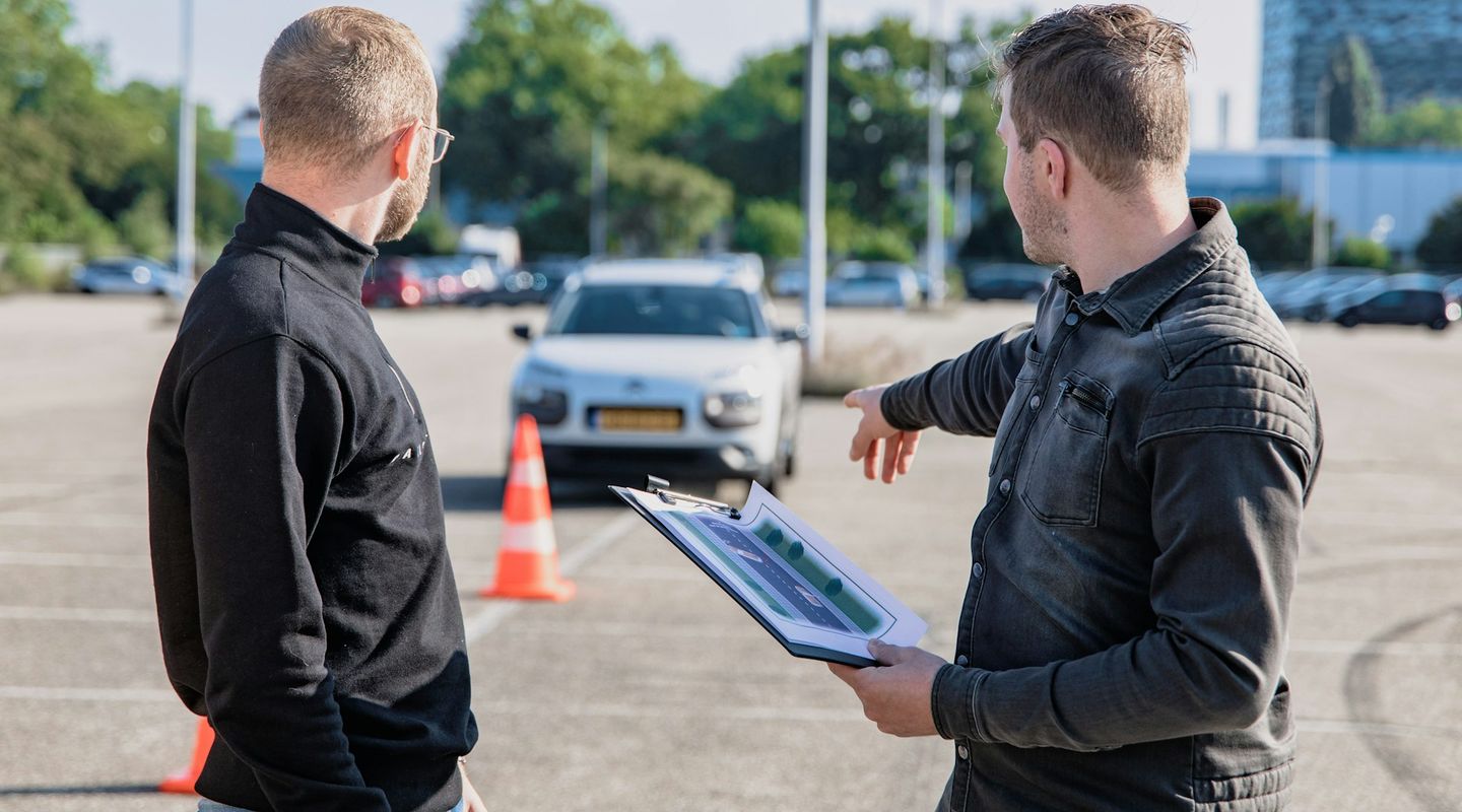 Driving instructor pointing and explaining to a student in an open car park with orange cones, a white car behind - driving w