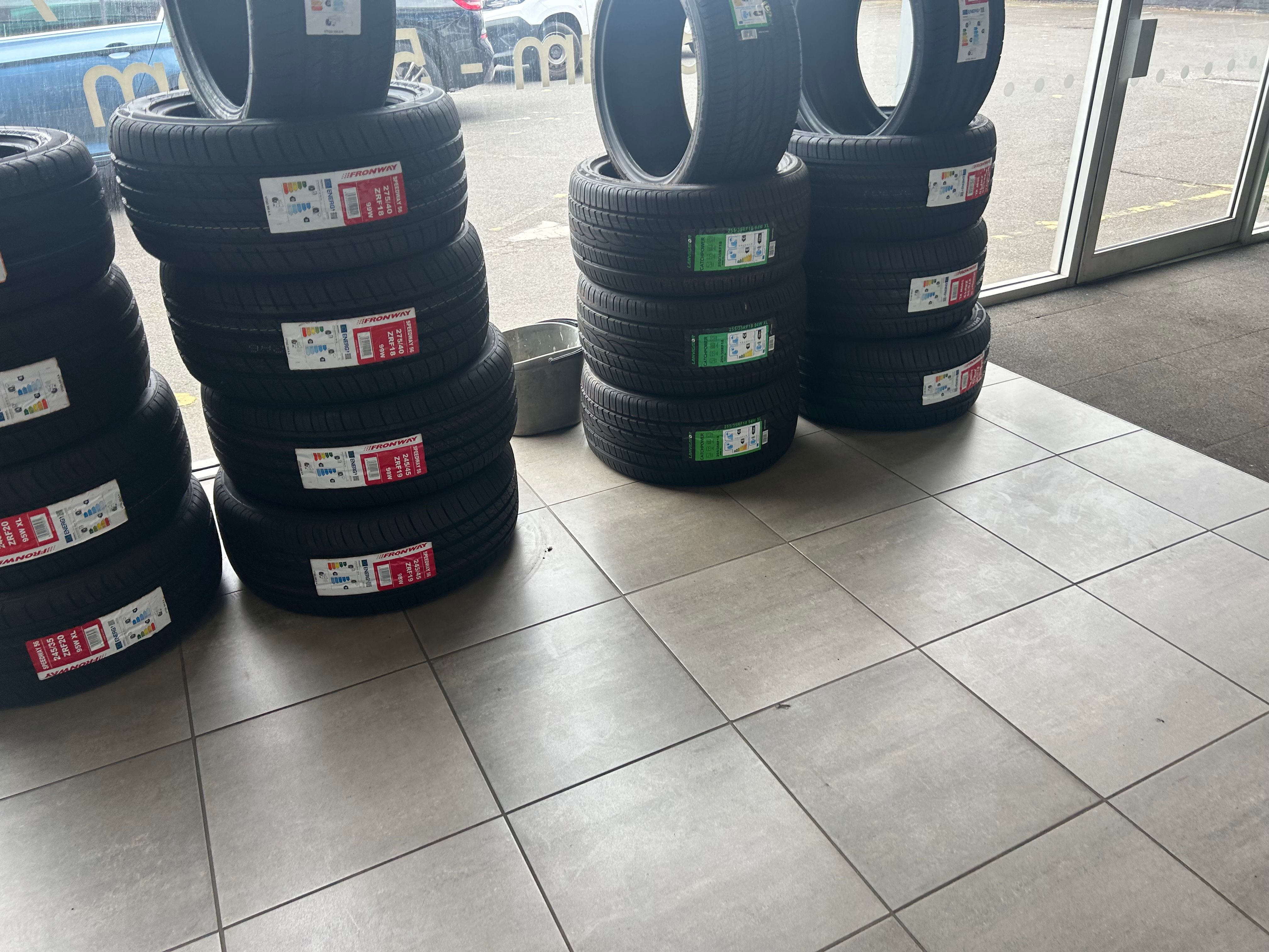 New branded tyres (Fronway and others) stacked on a garage floor, with a wet car park visible outside.