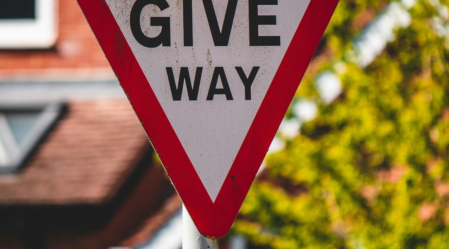Close-up of a UK "GIVE WAY" inverted triangular sign with red border, a Signature speed indicator mounted on top, bokeh of re