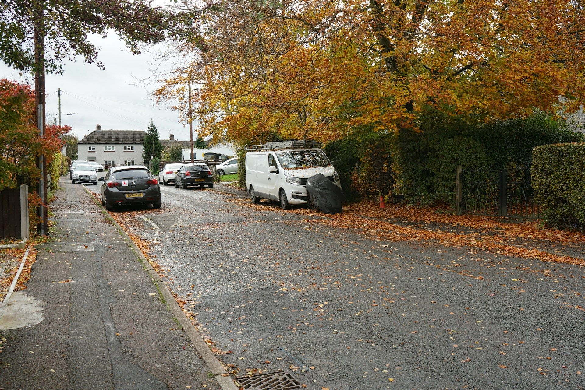 UK suburban street in autumn with fallen leaves on the road, a white transit van parked on the right and a car with L-plates