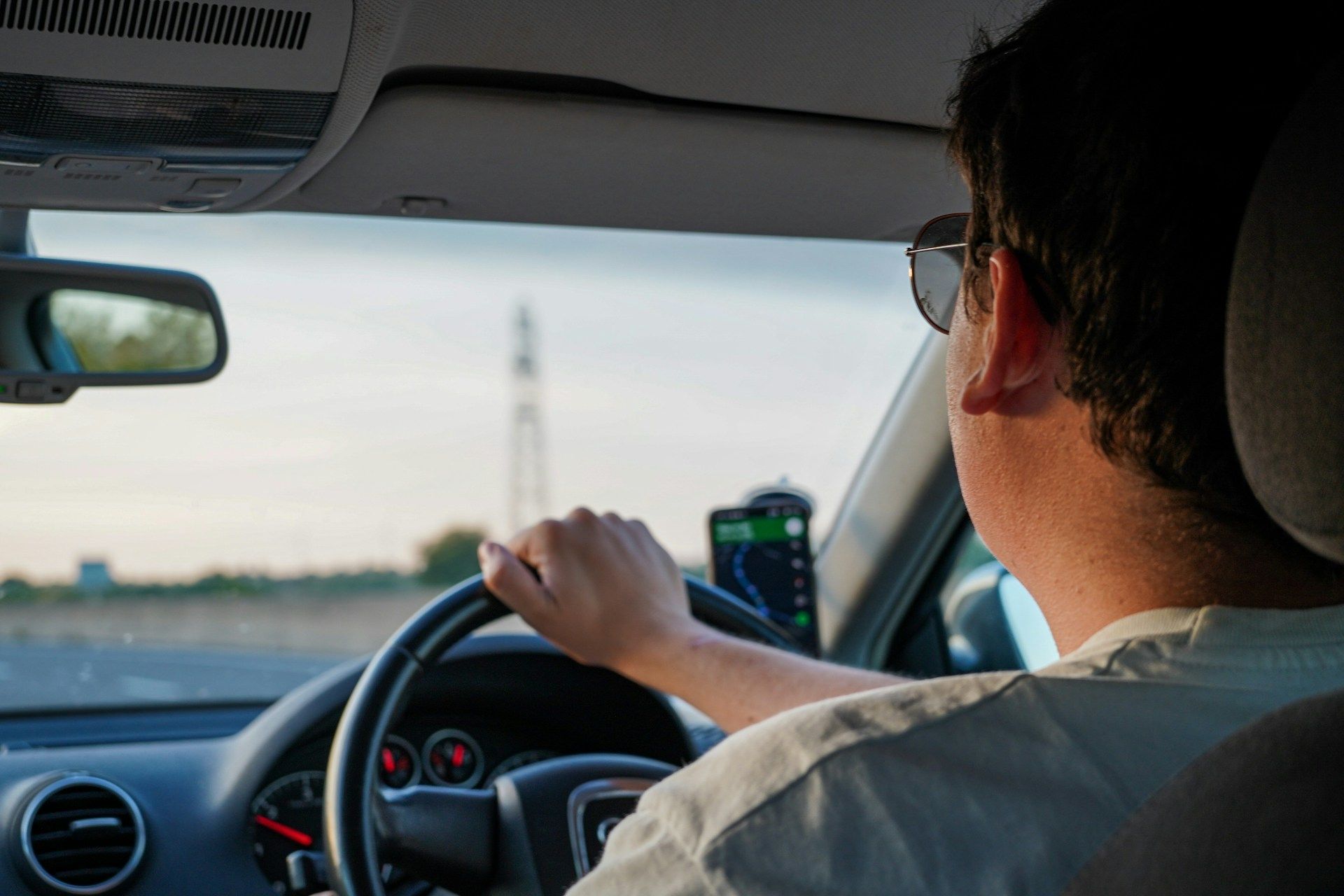 Driver wearing sunglasses at the wheel of a right-hand drive car with a phone satnav mount showing Google Maps, dusk light ou