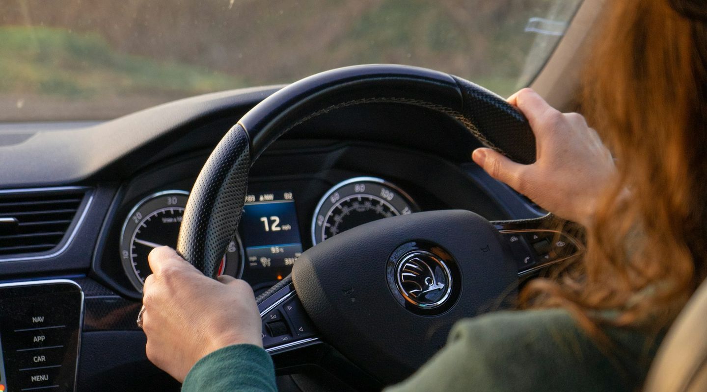 Woman with auburn hair at the wheel of a right-hand drive Skoda, golden winter light through bare trees outside - no claims d