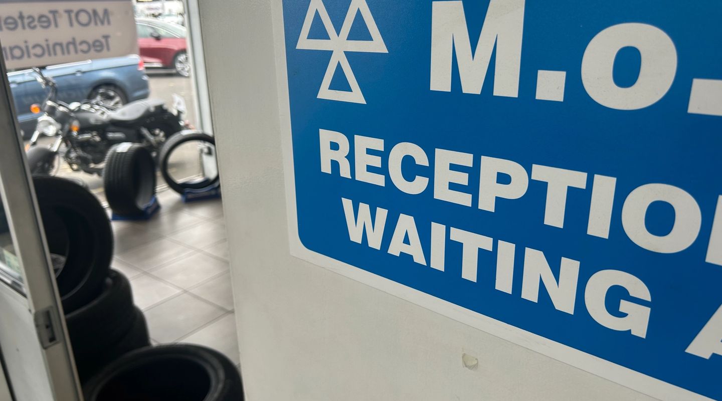 Close-up of a blue MOT Reception and Waiting Area sign, with tyres and a motorcycle visible in the background.