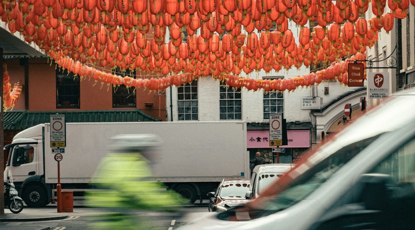 London Chinatown street with red lanterns overhead, white Ford Transit van blurred in the foreground, delivery lorry and cycl