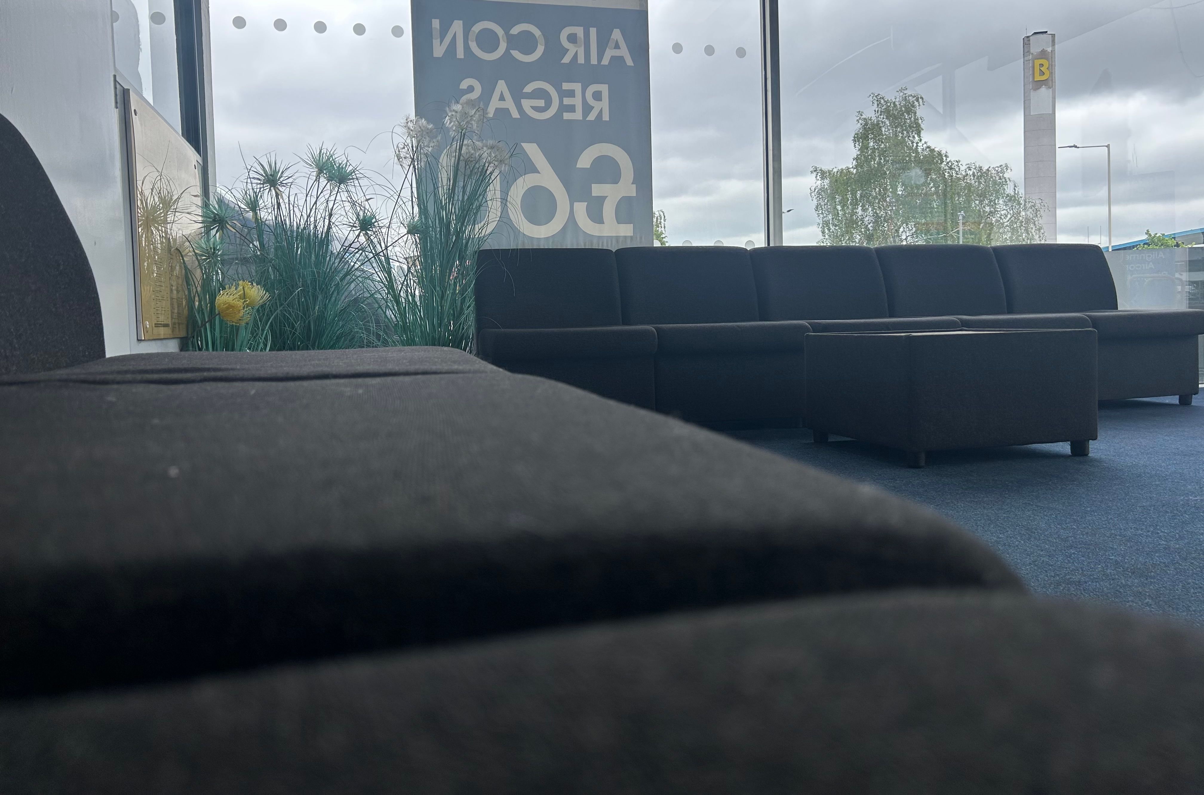 Low-angle view of a garage customer waiting room with dark sofas, plants, and a window showing the forecourt.