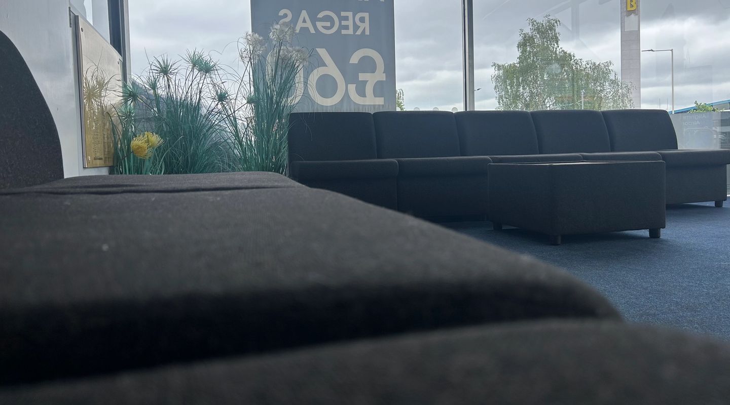 Low-angle view of a garage customer waiting room with dark sofas, plants, and a window showing the forecourt.