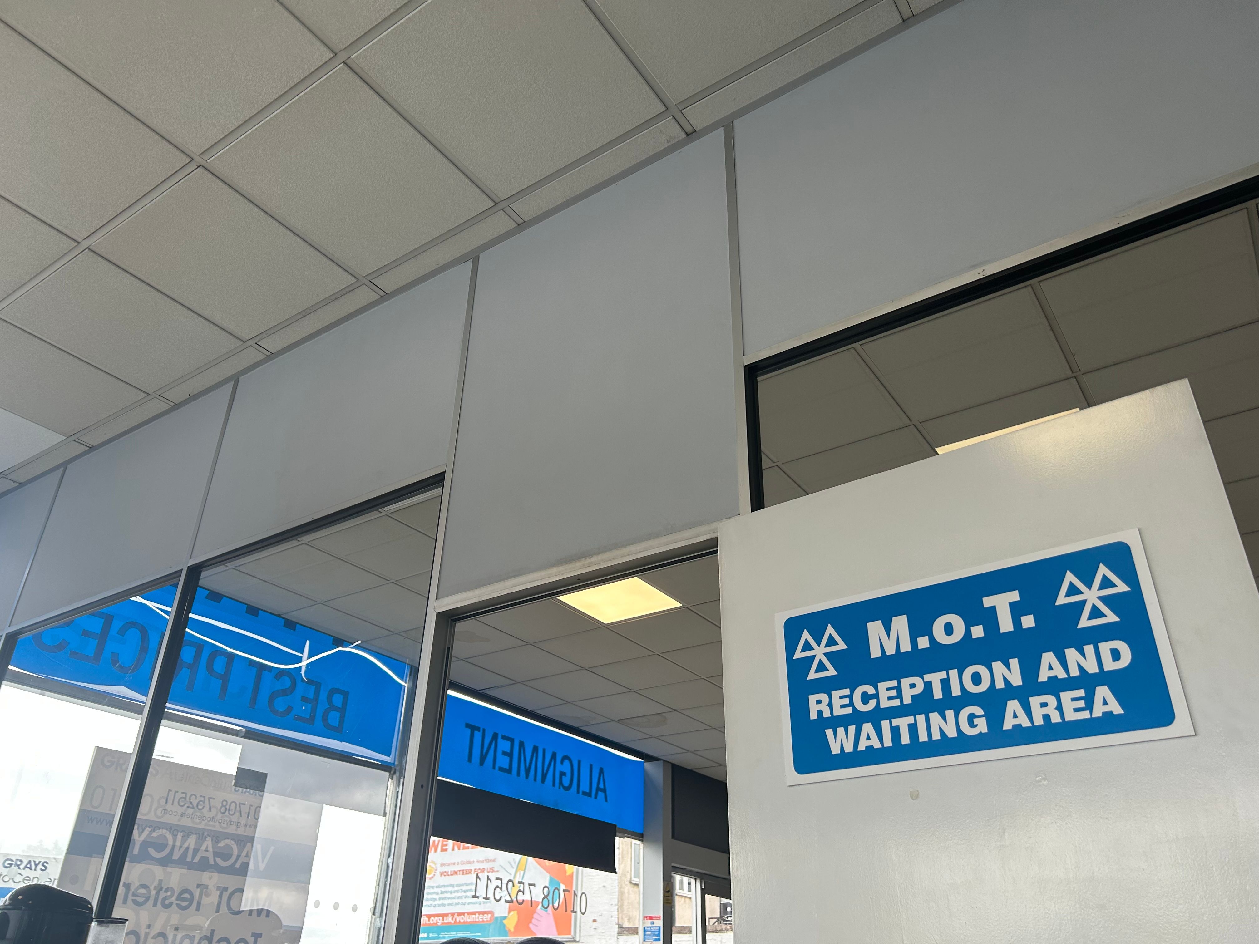 MOT M.o.T. Reception and Waiting Area sign on a wall, viewed from below with garage ceiling panels and windows.