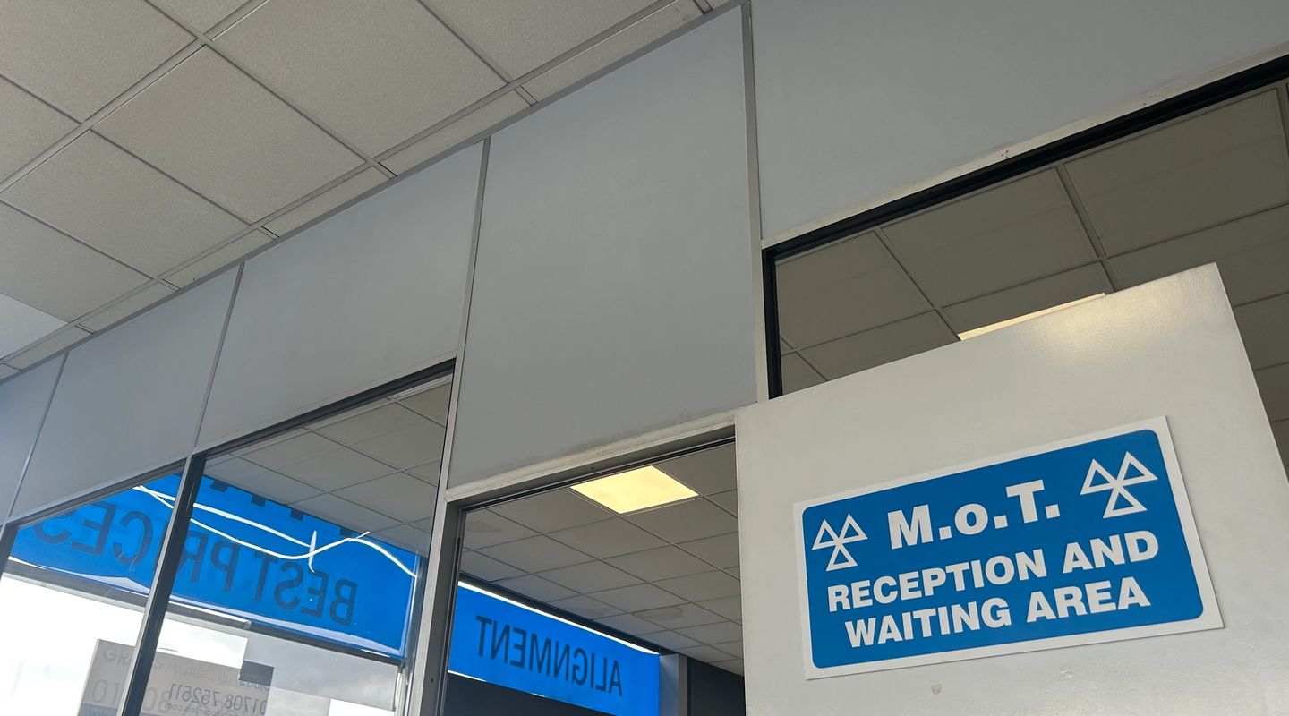 MOT M.o.T. Reception and Waiting Area sign on a wall, viewed from below with garage ceiling panels and windows.