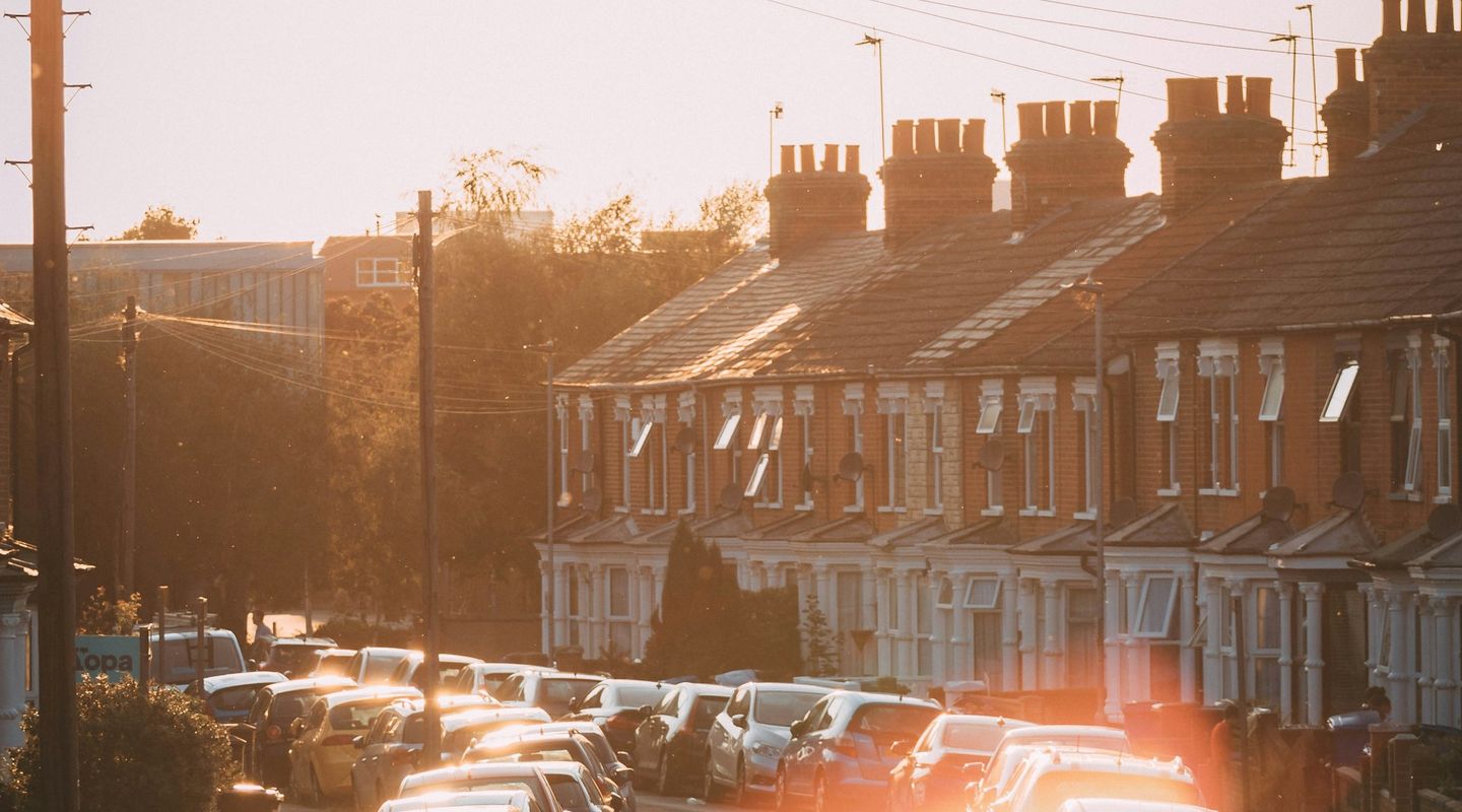 UK red-brick terraced street at golden hour, cars parked tightly on both sides, strong sun lens flare, chimney stacks silhoue