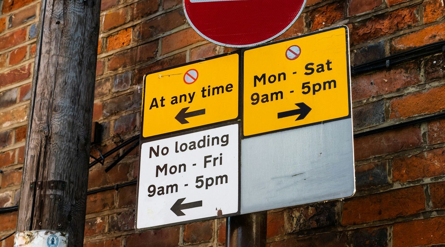 UK parking restriction signs: yellow-and-black "At any time" and "Mon-Sat 9am-5pm" direction panels with a no-entry circle ab