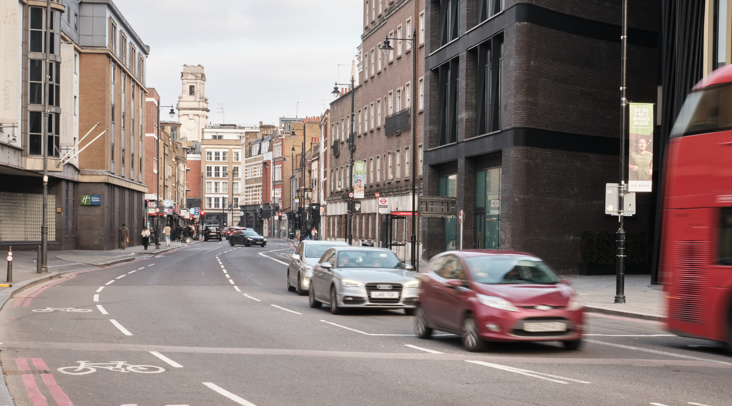 Mid-day London traffic on a wide road, red Ford Fiesta and silver Audi A3 in motion, a passing red bus on the right.