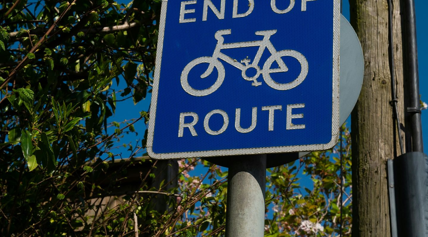 Blue "END OF ROUTE" cycle route sign with bicycle pictogram against a blue sky with green foliage - cycle lanes uk