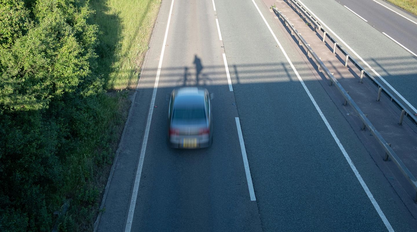 UK dual carriageway viewed from a bridge overhead, a car passing below with the photographer's shadow cast on the tarmac, gre