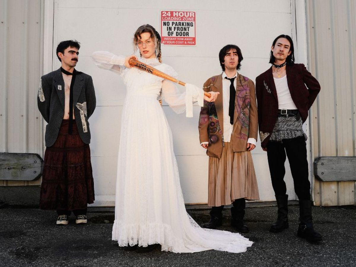 Four members of PISS pose outside an industrial building, wearing eclectic, punk-inspired outfits; one member stands in a long white dress holding a translucent prop across their shoulder.