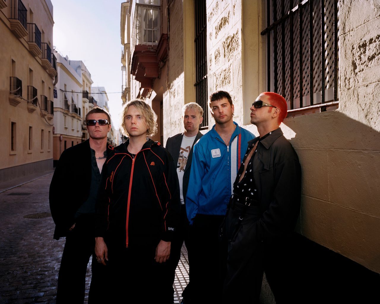Five members of Fontaines D.C. pose beside a sunlit wall on a narrow cobblestone street.
