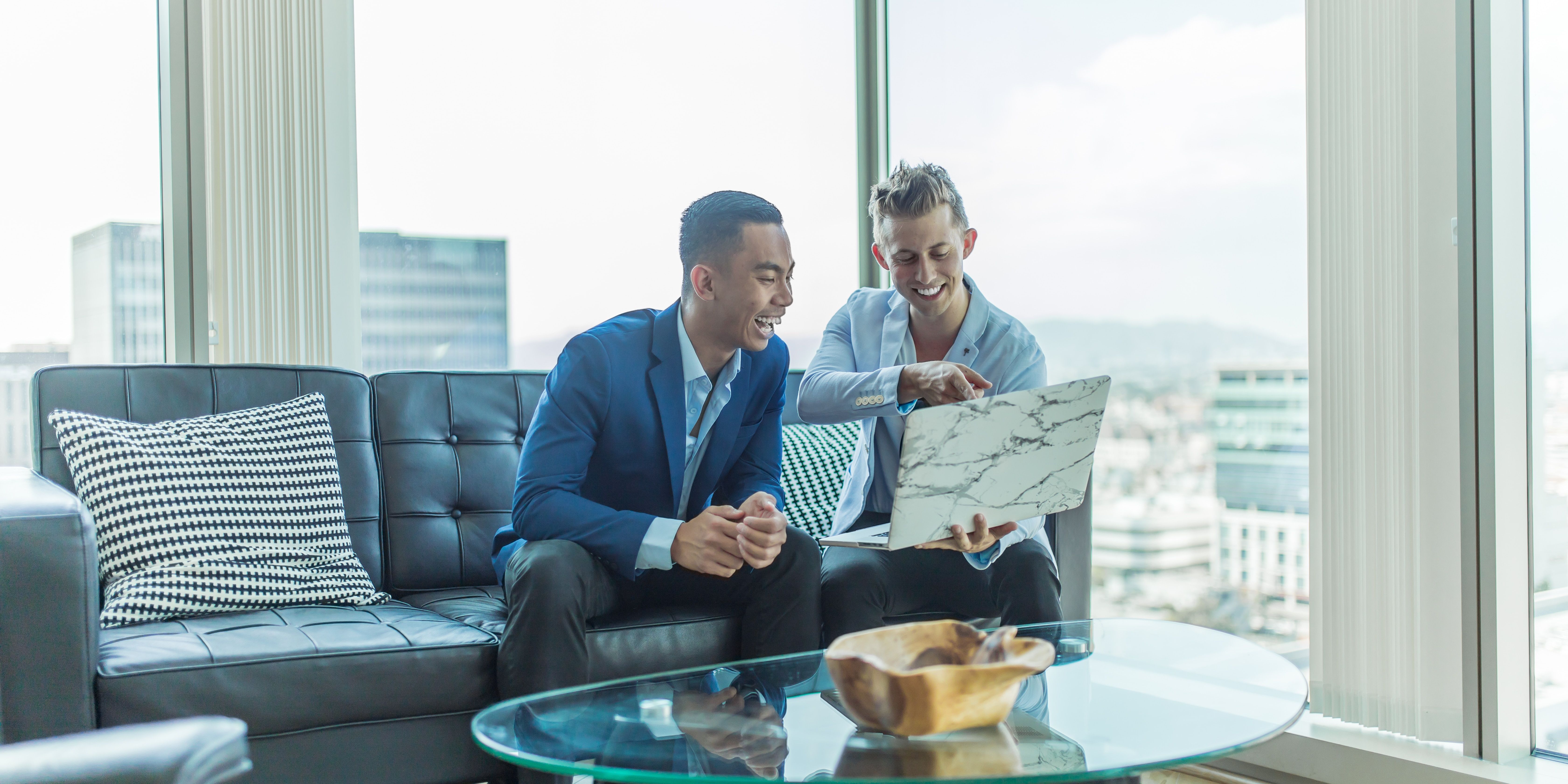Two accountants laugh near a large window in a skyscraper, while perusing a laptop together
