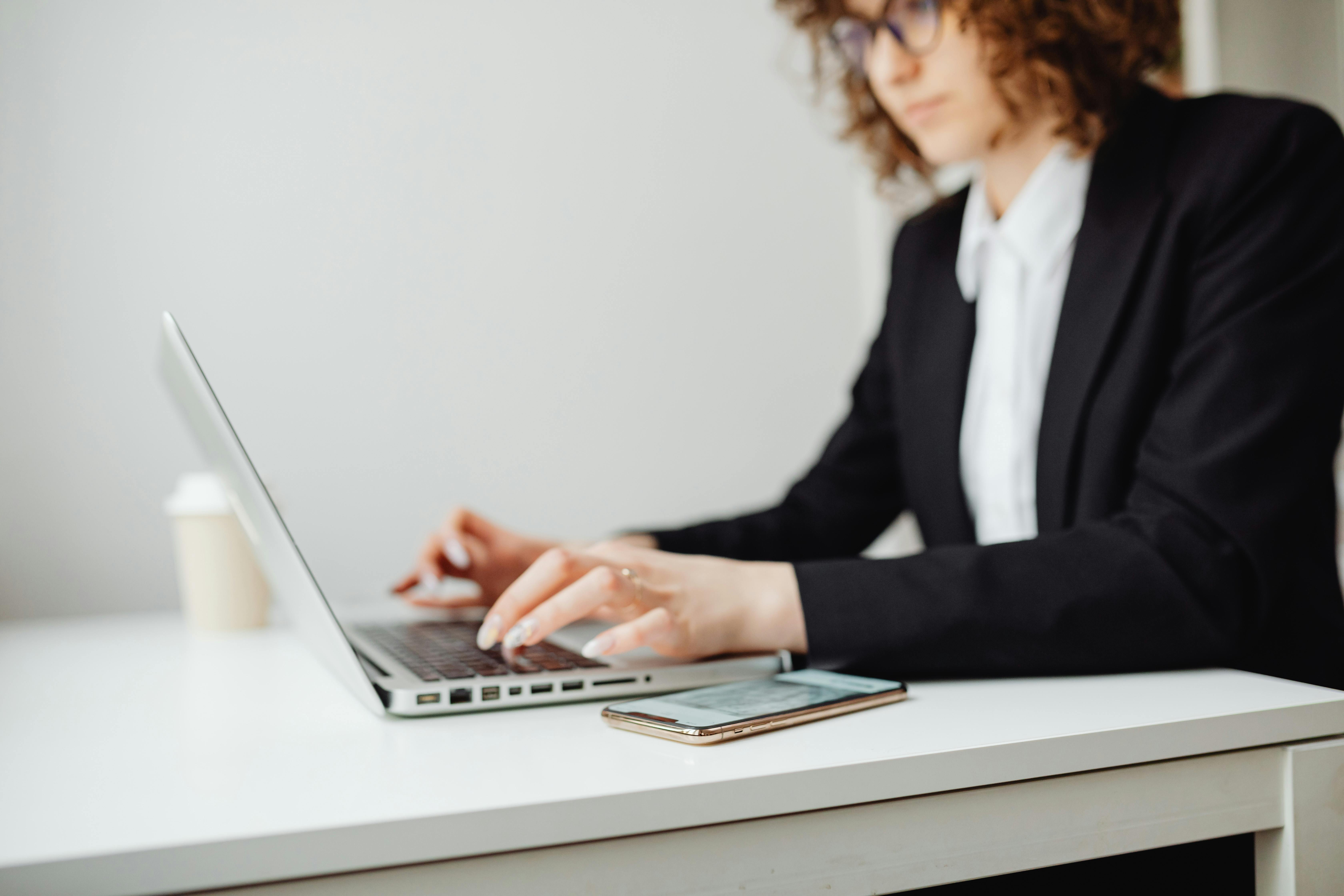 A professional woman in a black blazer and white shirt is typing on a laptop at a white desk. A smartphone and a coffee cup are nearby. She has curly hair and wears glasses, appearing focused on her work.