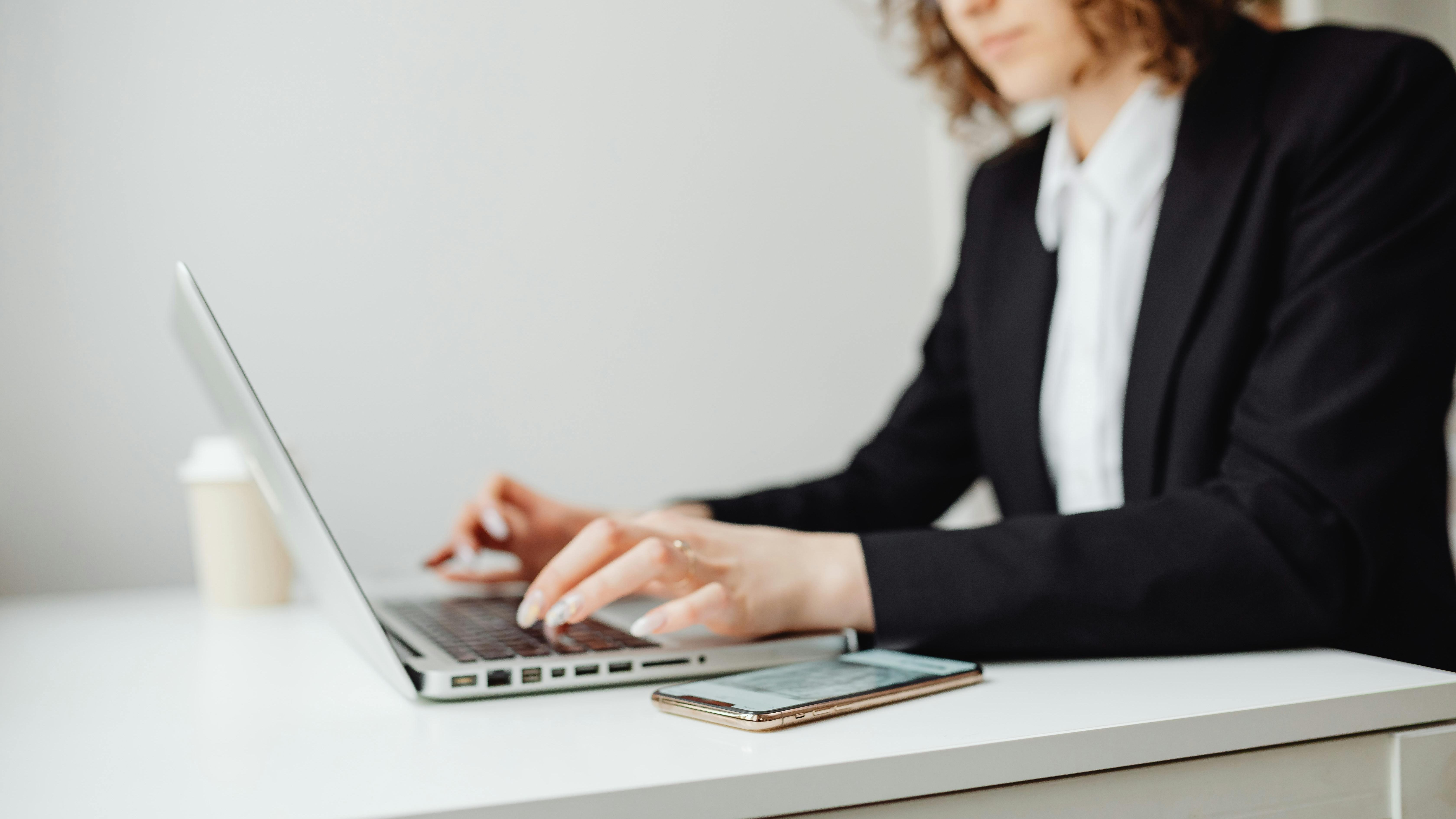 A professional woman in a black blazer and white shirt is typing on a laptop at a white desk. A smartphone and a coffee cup are nearby. She has curly hair and wears glasses, appearing focused on her work.