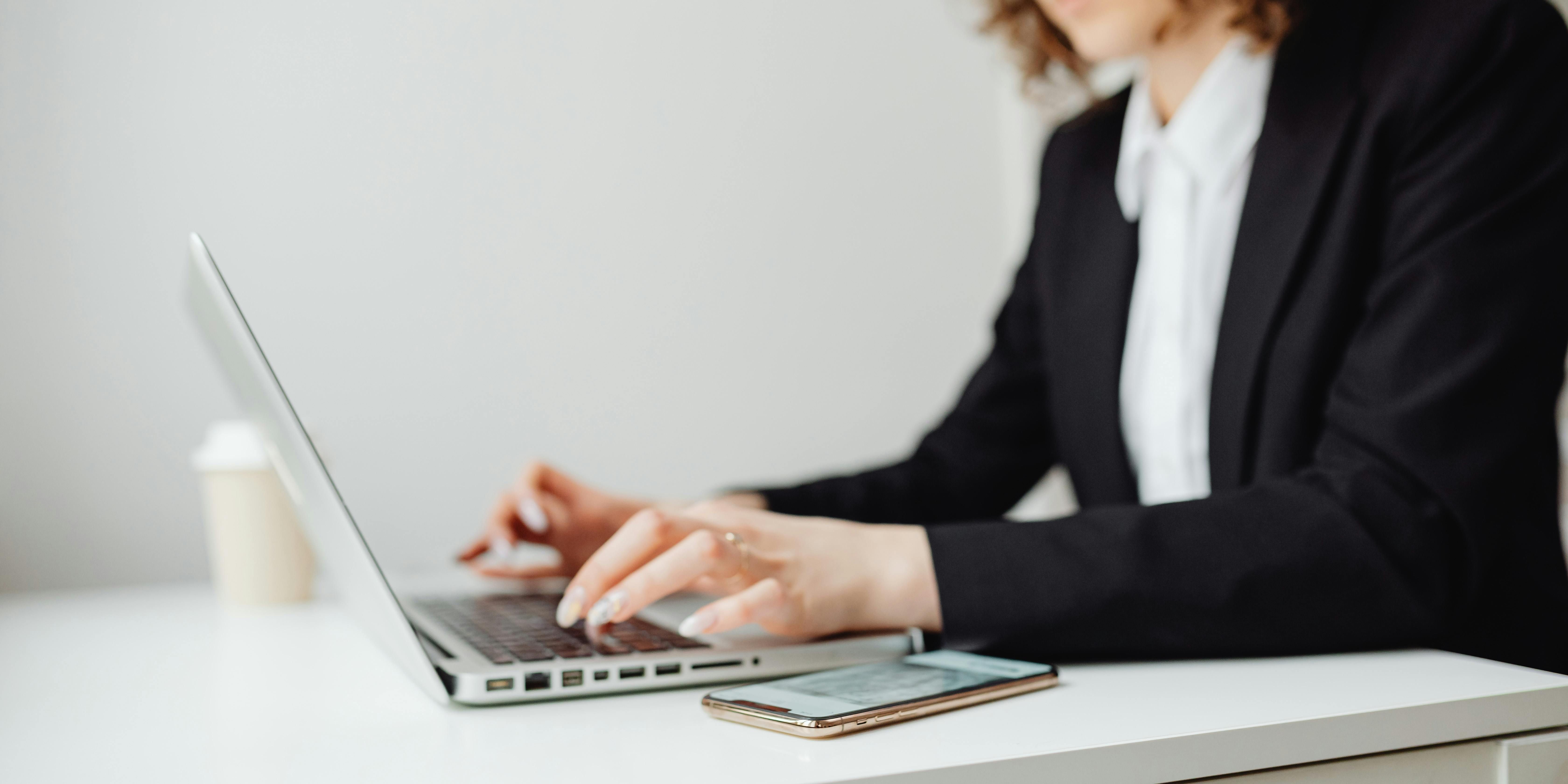 A professional woman in a black blazer and white shirt is typing on a laptop at a white desk. A smartphone and a coffee cup are nearby. She has curly hair and wears glasses, appearing focused on her work.