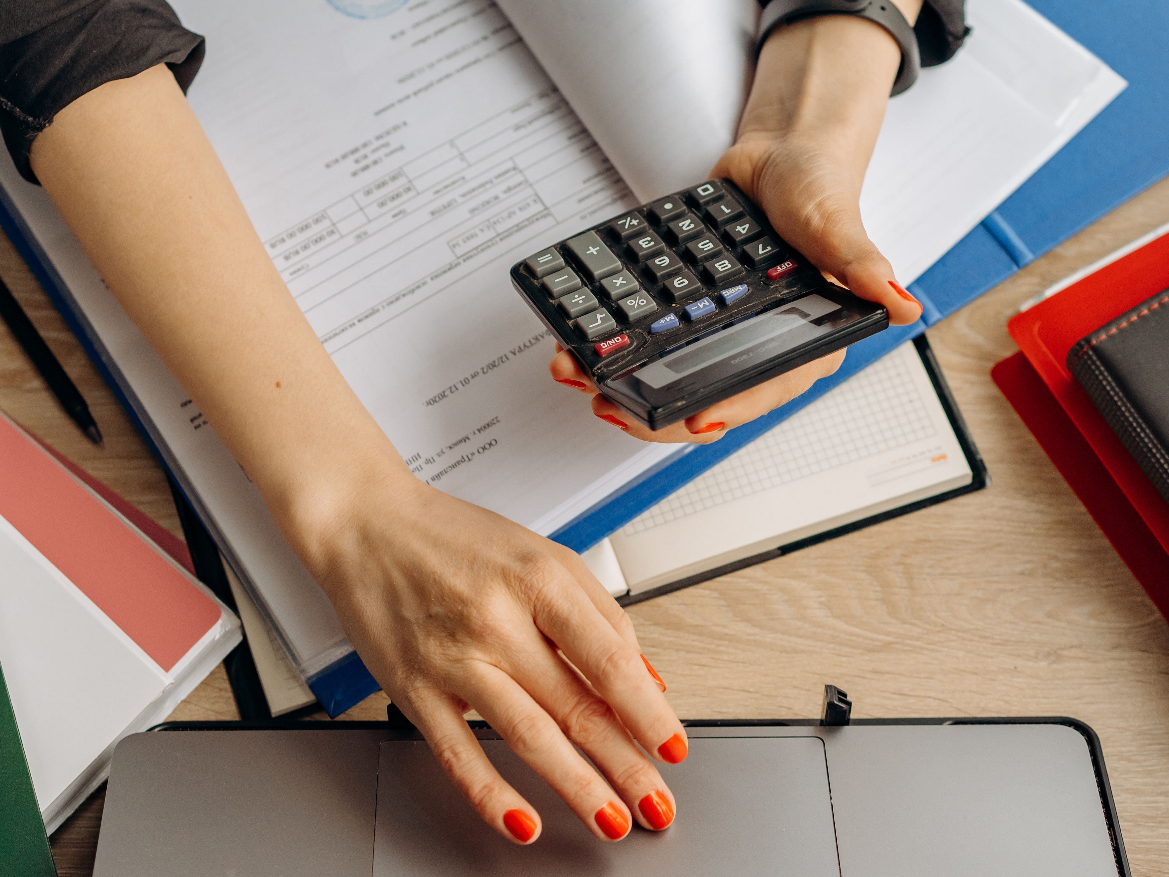 A person with orange nails uses a calculator in one hand and a laptop with the other.