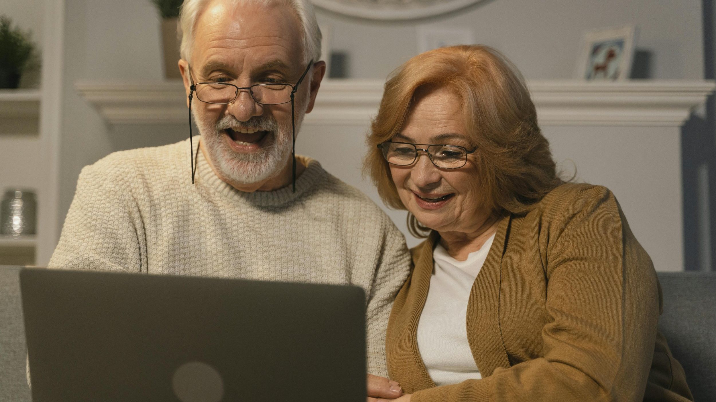 An elderly couple with glasses is sitting together on a couch, smiling while looking at a laptop screen. The man, wearing a beige sweater, appears excited, while the woman in a brown cardigan looks happy and engaged.
