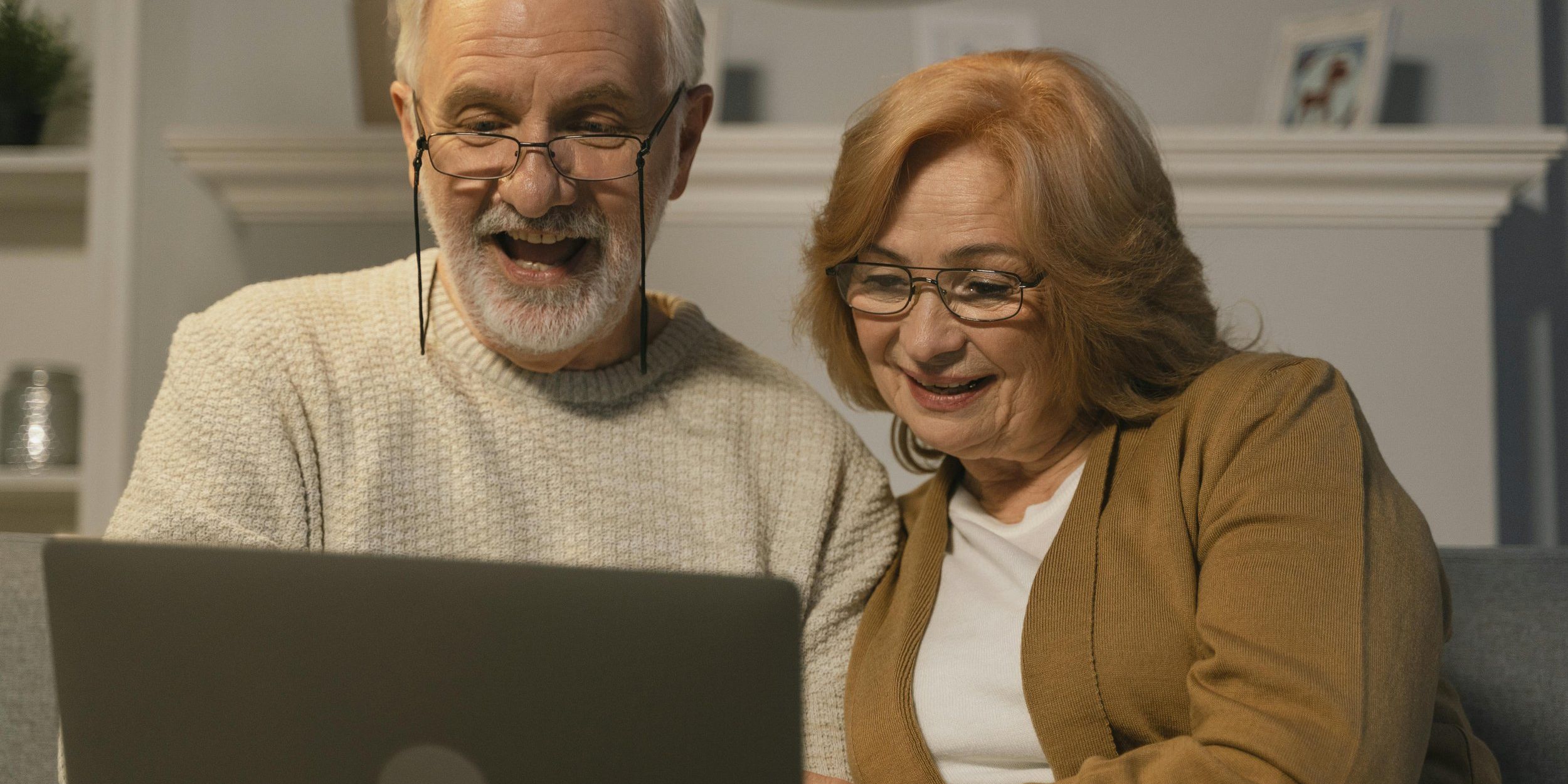 An elderly couple with glasses is sitting together on a couch, smiling while looking at a laptop screen. The man, wearing a beige sweater, appears excited, while the woman in a brown cardigan looks happy and engaged.
