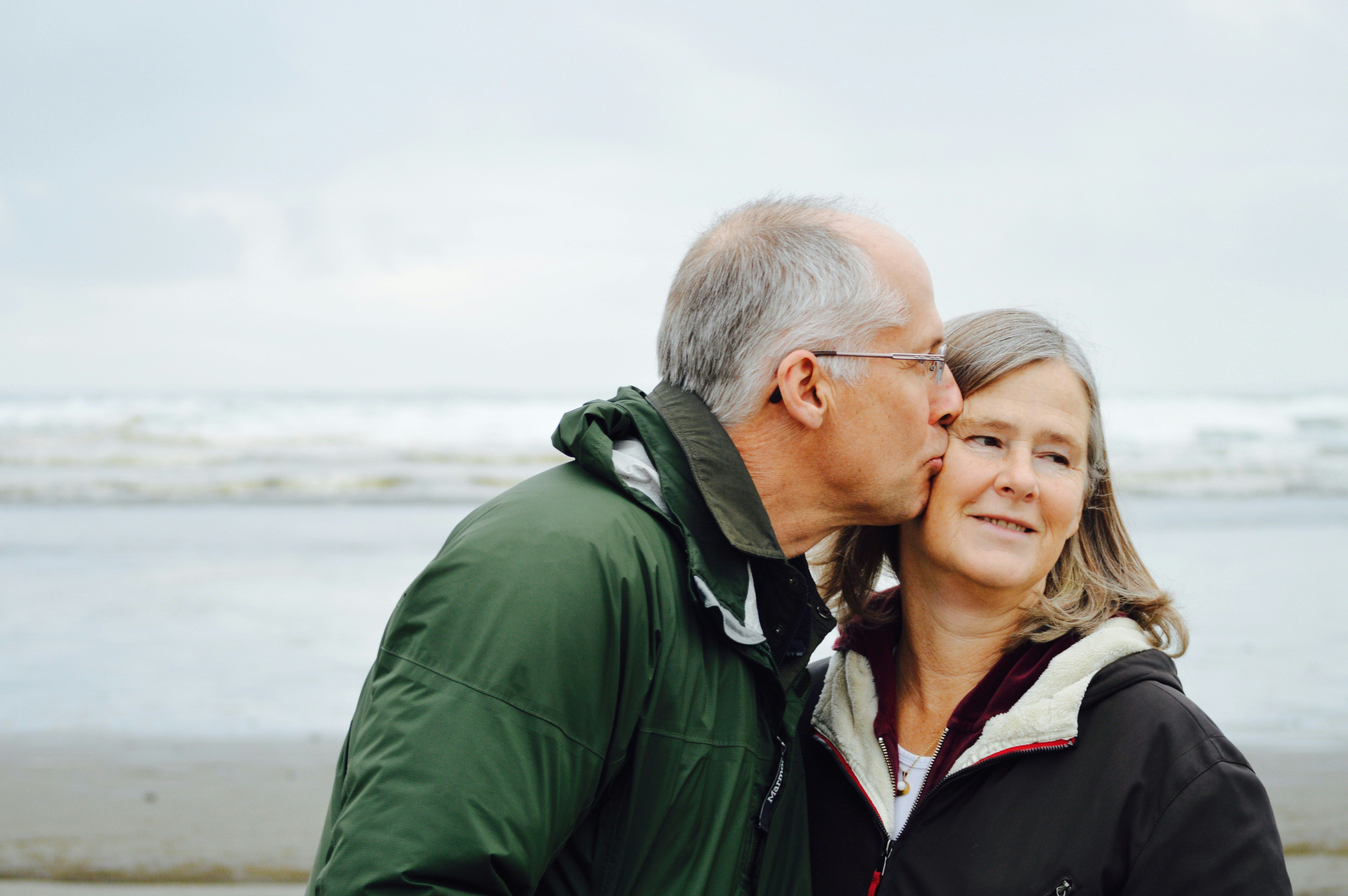 A man with grey hair kisses the cheek of a woman while they stand on a beach.