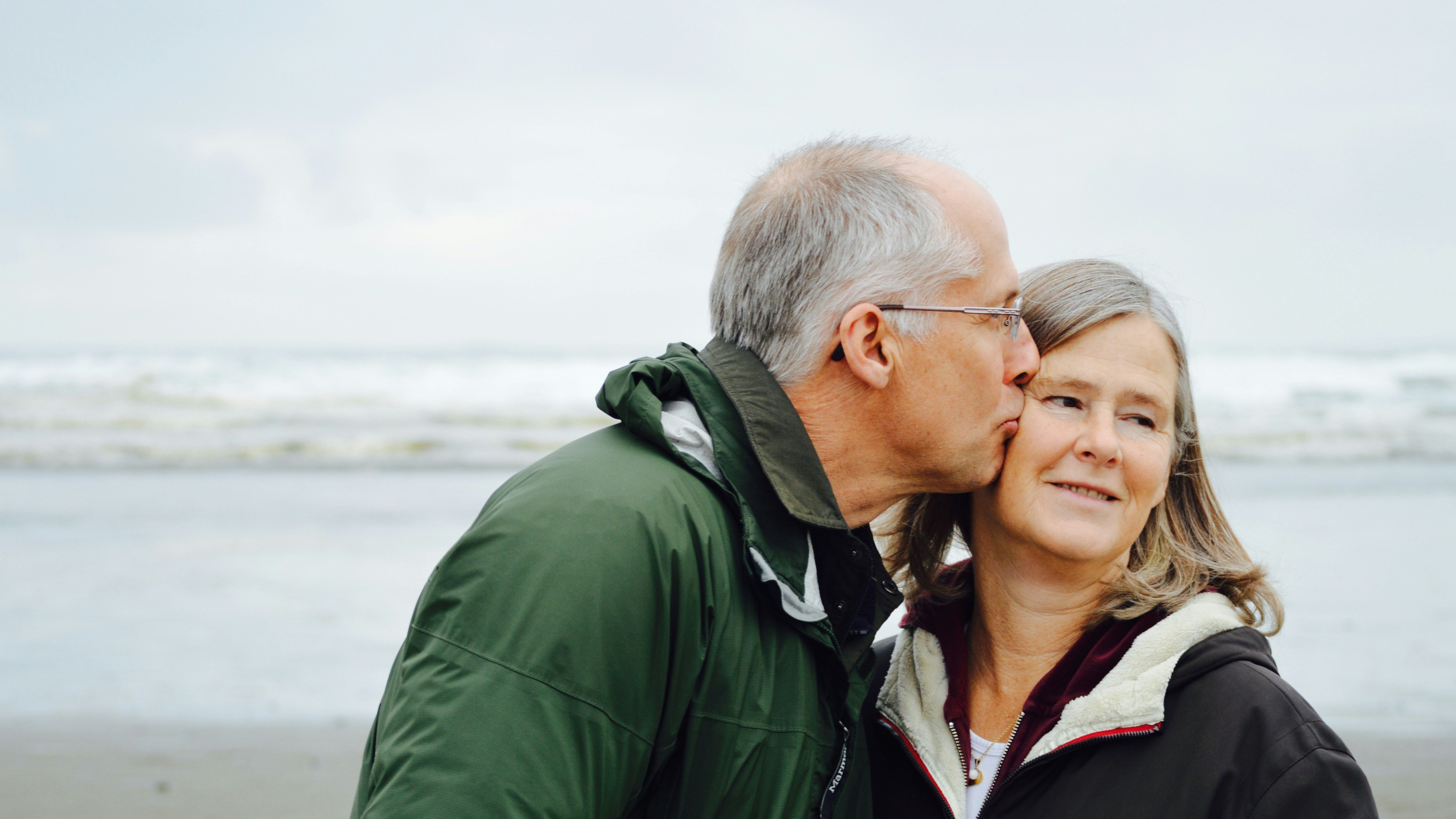 A man with grey hair kisses the cheek of a woman while they stand on a beach.