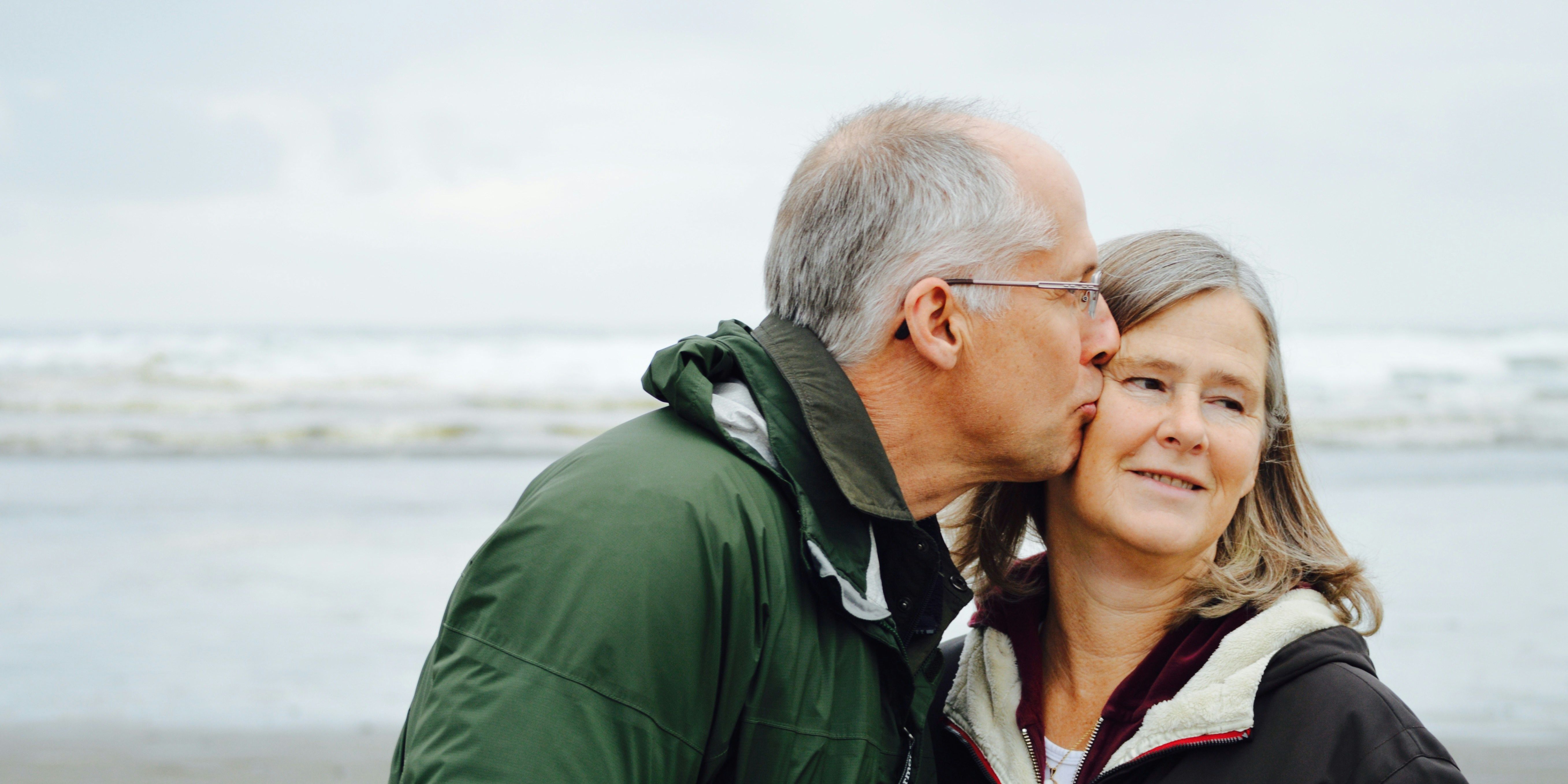 A man with grey hair kisses the cheek of a woman while they stand on a beach.