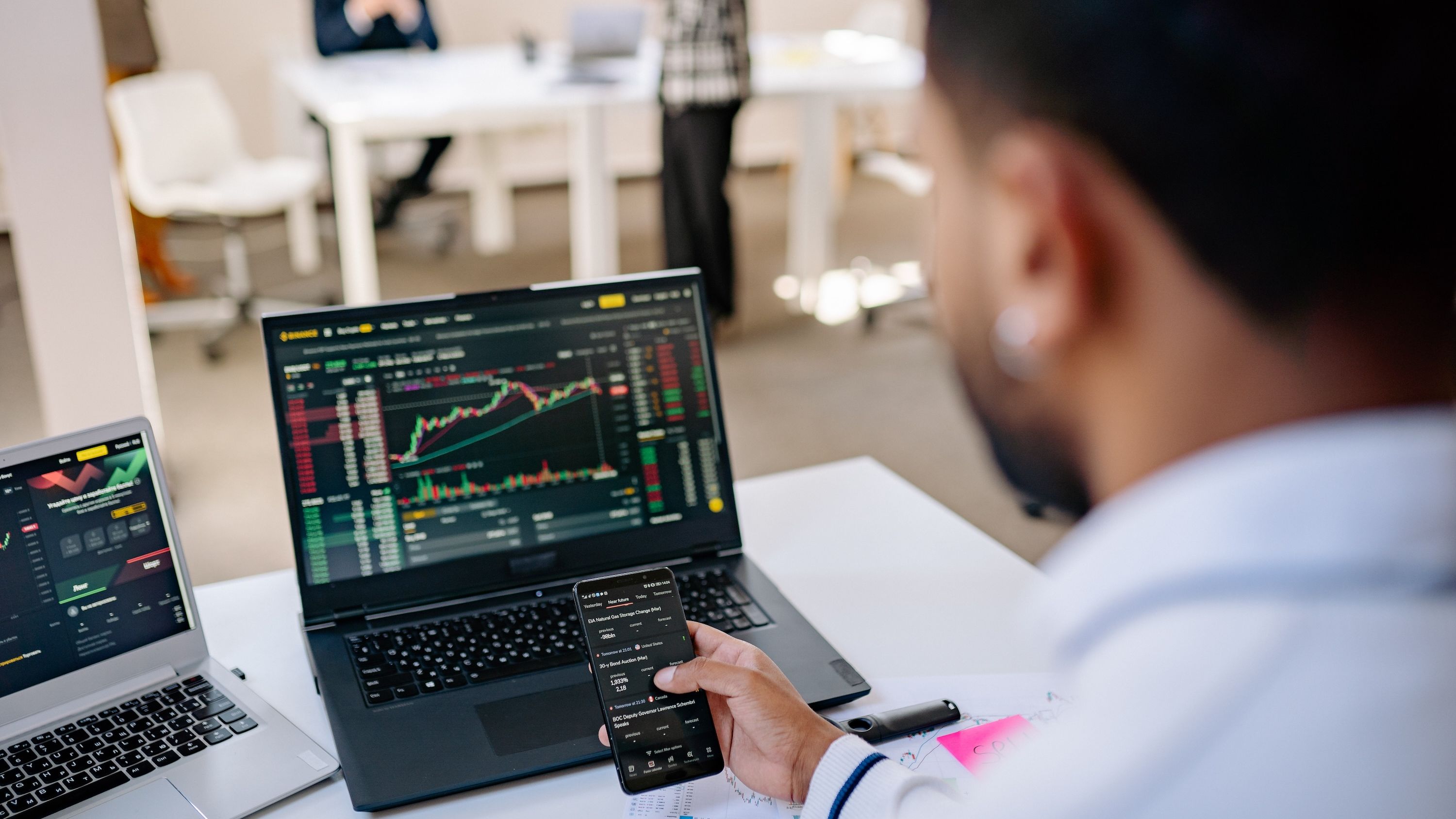 A man flips through an app on his phone, while perusing graphs on a laptop behind the phone.